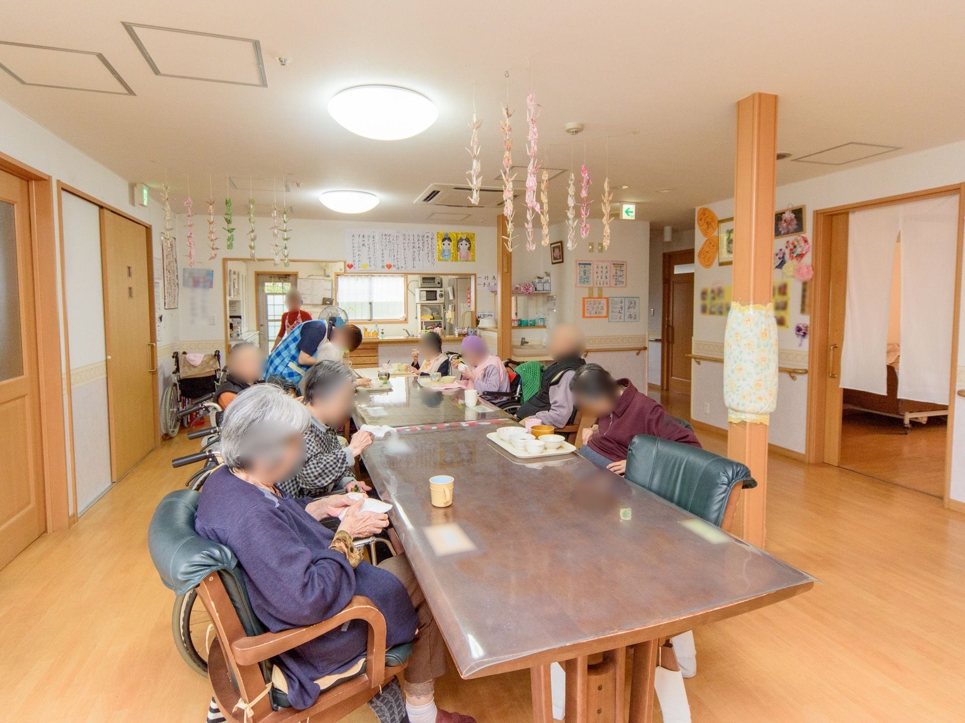 テーブルセットが２つ並べられたフロアに入居者が集まっている。スタッフに見守られながら、食事をしたりお茶を飲んだりしている。