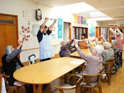 スタッフが立ち、高齢者たちは座ったままで帽を両手で持って上に挙げる運動をしている。