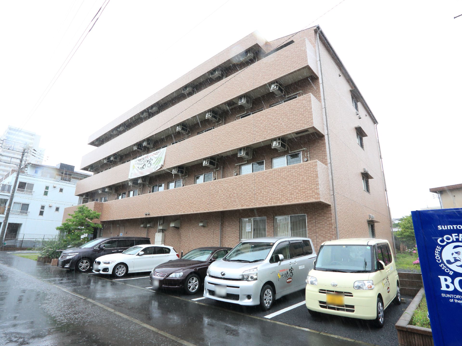施設名が書かれた垂れ幕が2階部分にある。建物の前には、6台分の駐車場がある。近くには自動販売機がある。