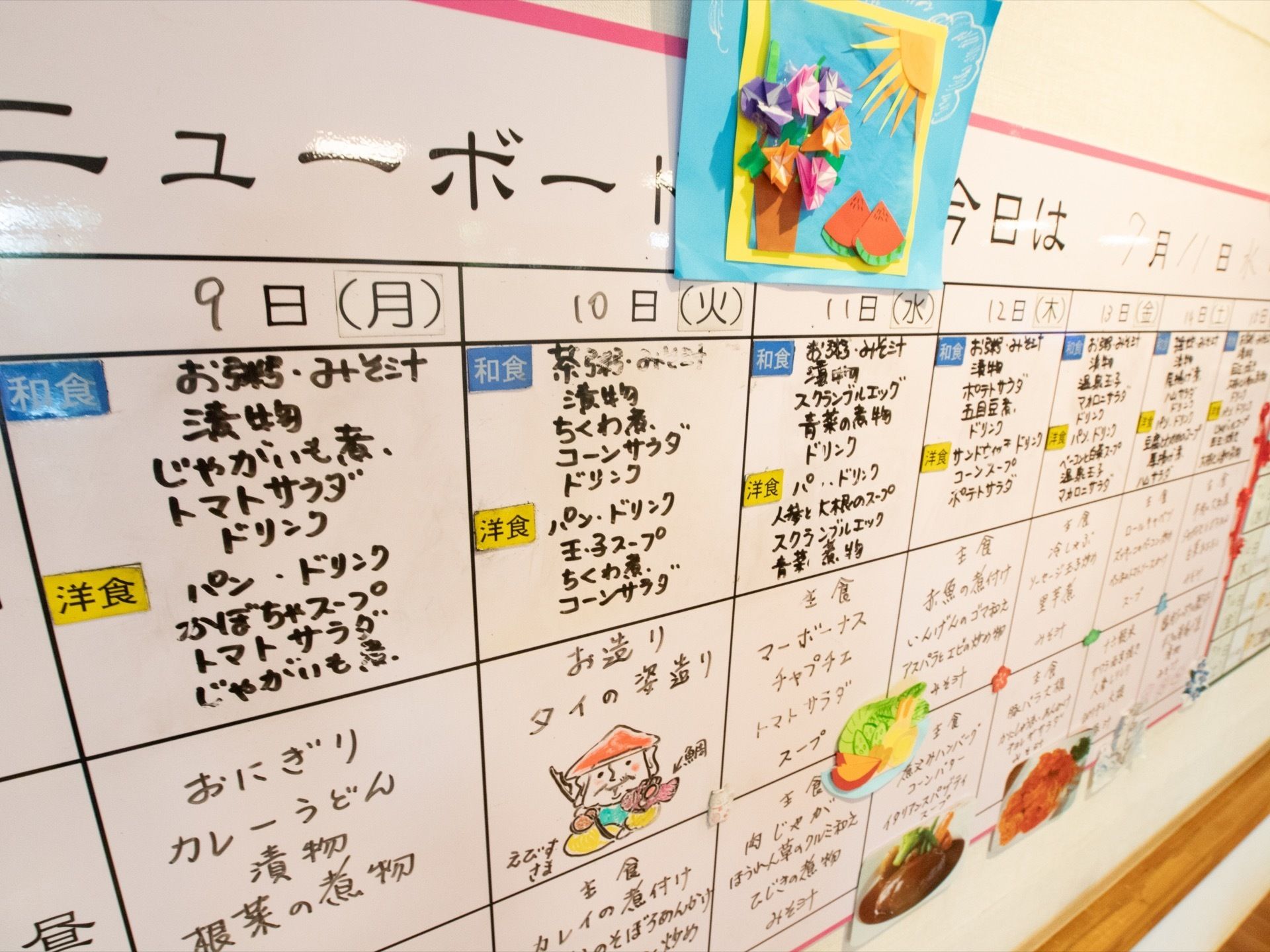 壁に食事のメニューが書かれたボードが貼られている。約１週間のメニューがまとめて書かれ、イラストなどが添えられている。