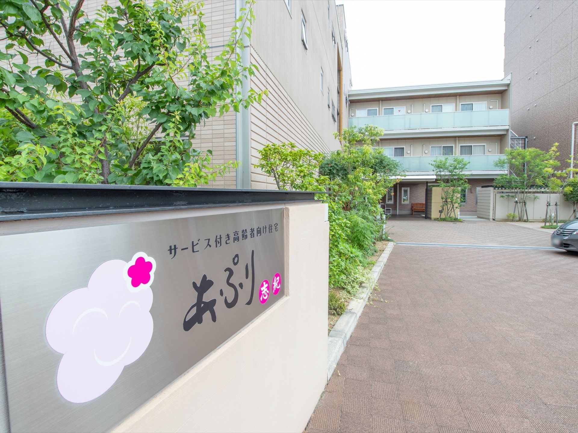 施設敷地入口にステンレス製の看板を取りつけている。建物前には樹木が植えられ、駐車スペースが用意されている。