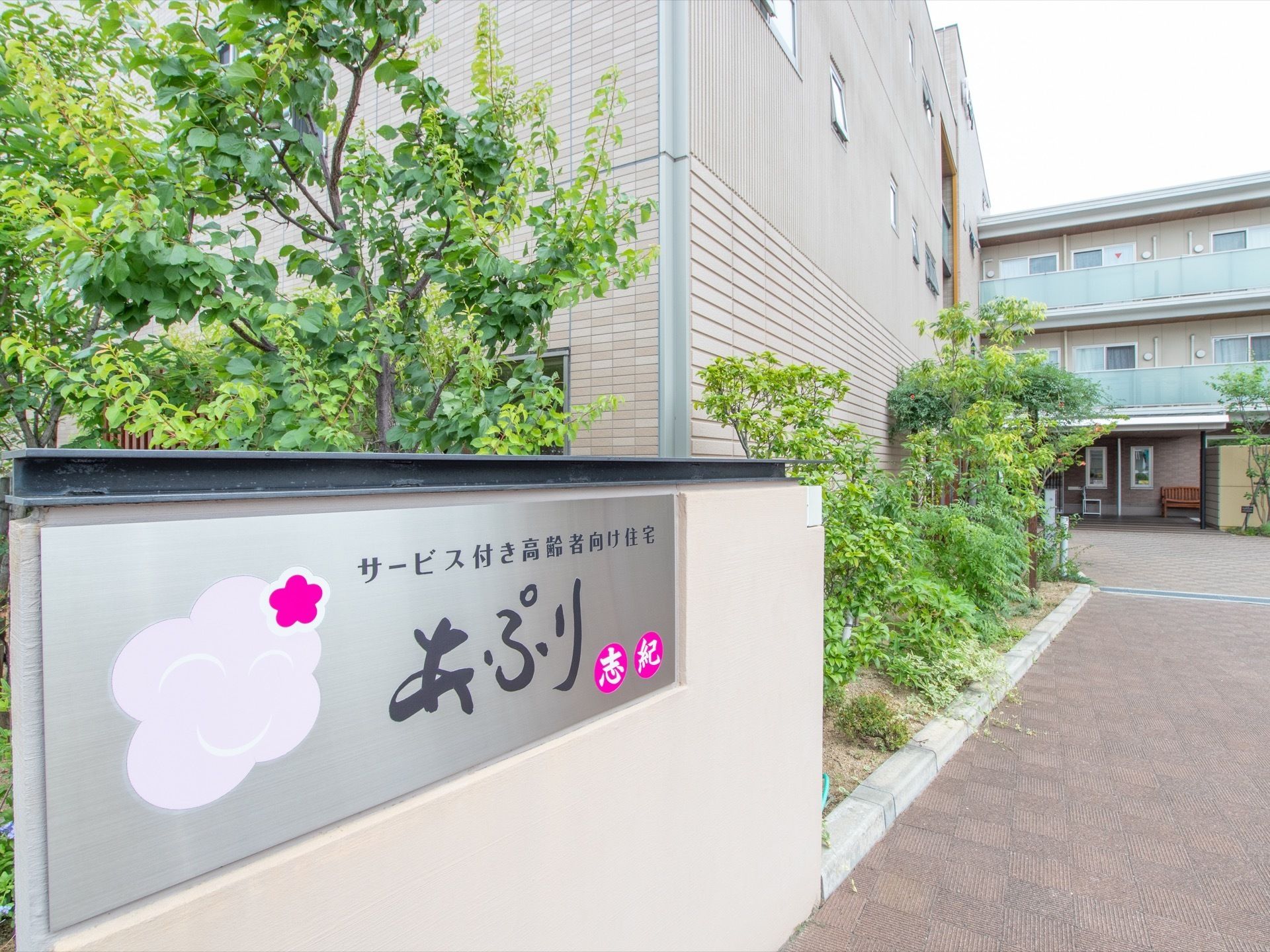 木々に囲まれた施設入り口。看板には施設名と共にあたたかみあるマークがある。どの個室にもベランダや窓が完備されている。