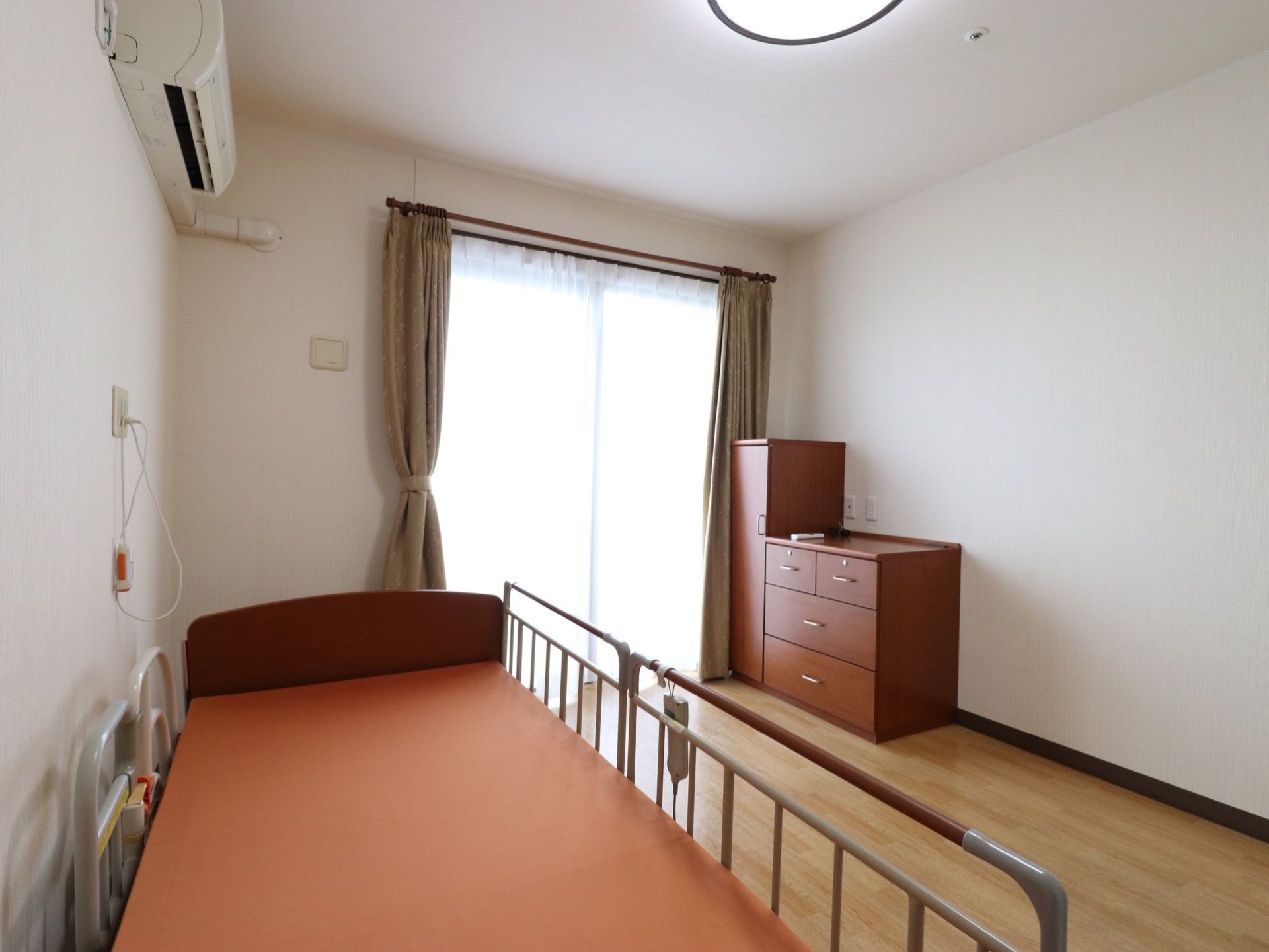 居室には電動ベッドが1台置かれている。洋服ダンスが2種類置かれ、エアコンがついている。大きな窓があり、フローリングで、ナースコールもある。