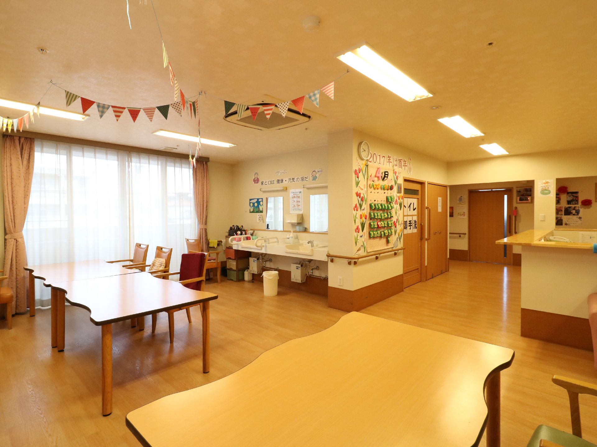 机とテーブル、洗面台がある。壁には手摺が設置されている。手作りの大きなカレンダーが飾られている。入口は扉が無く広い。