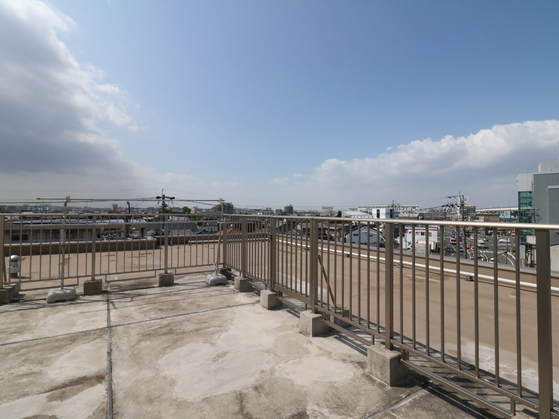 広くて開放的な屋上。物干竿が置かれている。周辺は大きな建物が少なく、遠くまで見通す事が出来る。フェンスから建物の淵まで距離がある。