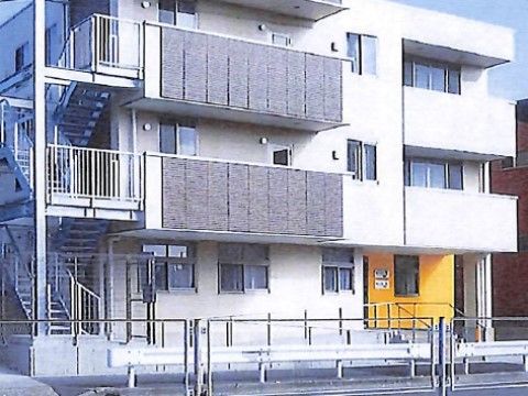 白い３階建ての建物に、金属製の外付け階段が設置されている。１階の一部が黄色い壁になっていて窓が取り付けられている。