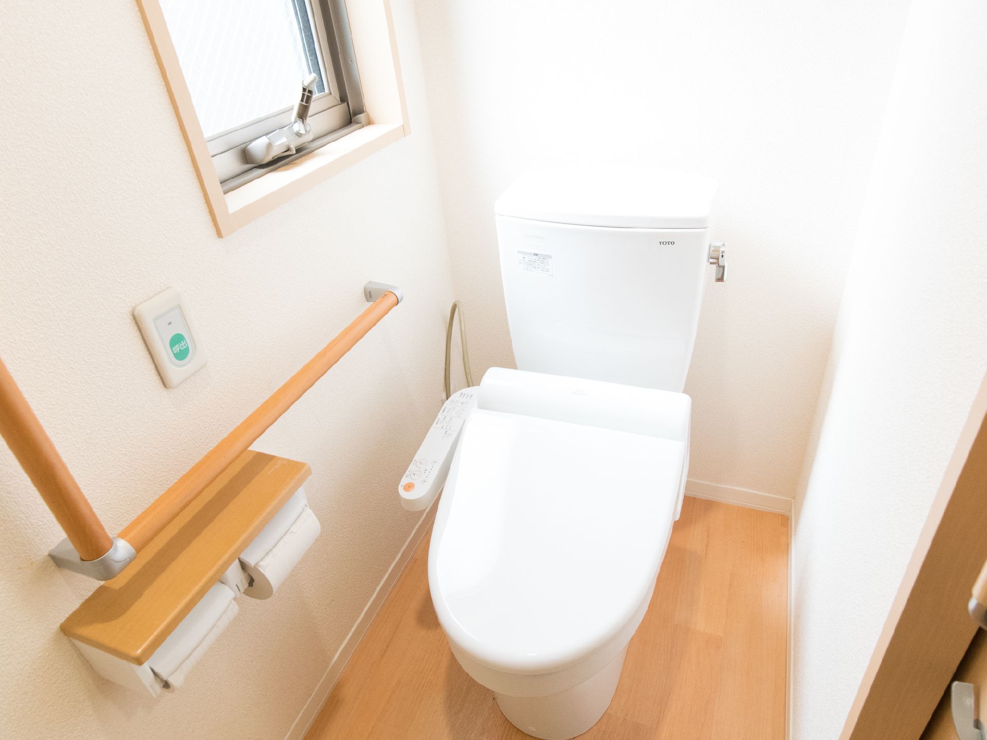 窓があり明るいトイレ。壁にL字型の手摺、トイレットペーバーのフォルダーが2つ、緊急呼出ボタンが設置されている。