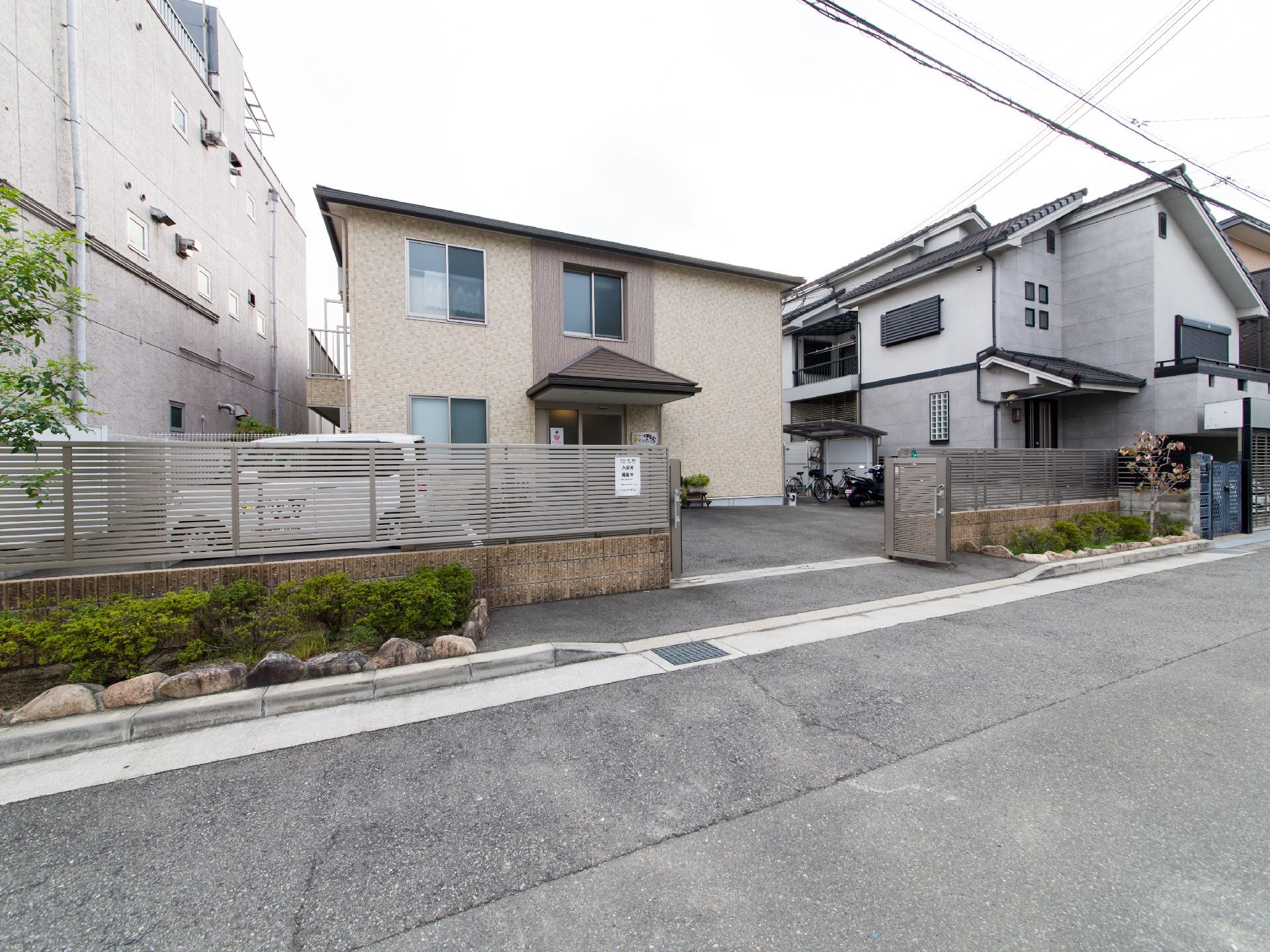 施設建物をフェンスの外、道路側から見たところ。正面ゲートの左右には石で囲まれた植え込みが作られている。