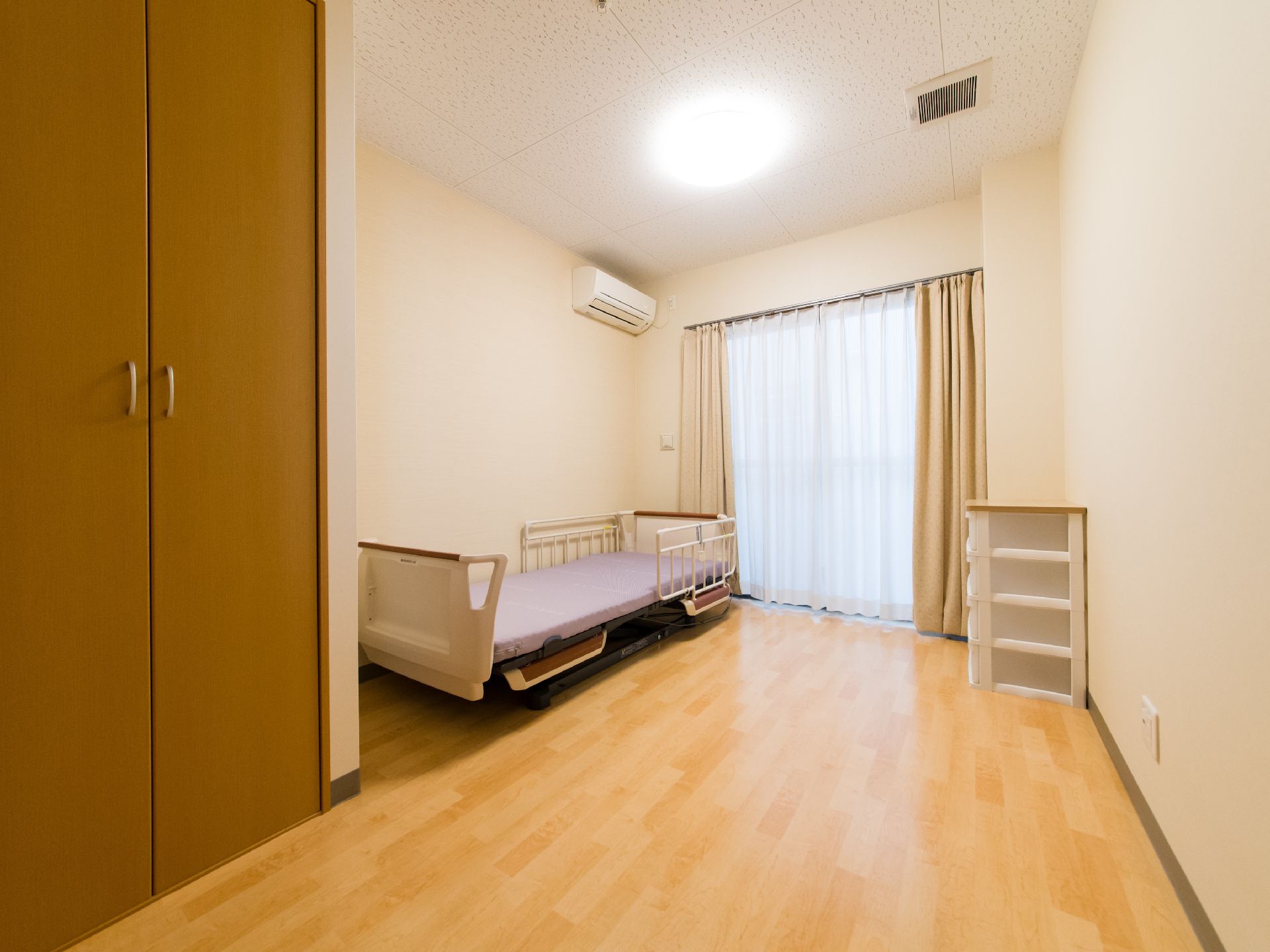 入居者様用の居室。壁と天井は白く、窓はベランダになっている。介護ベッドの真上にはエアコンが設置されている。