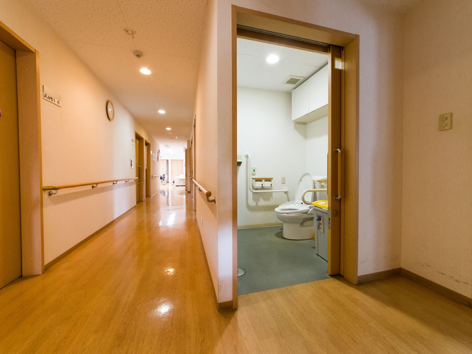 廊下の奥には、ソファが置いてあるのが見える。車いすの人がいるのが見える。写真の右手前に、洋式トイレがあるのが見える。