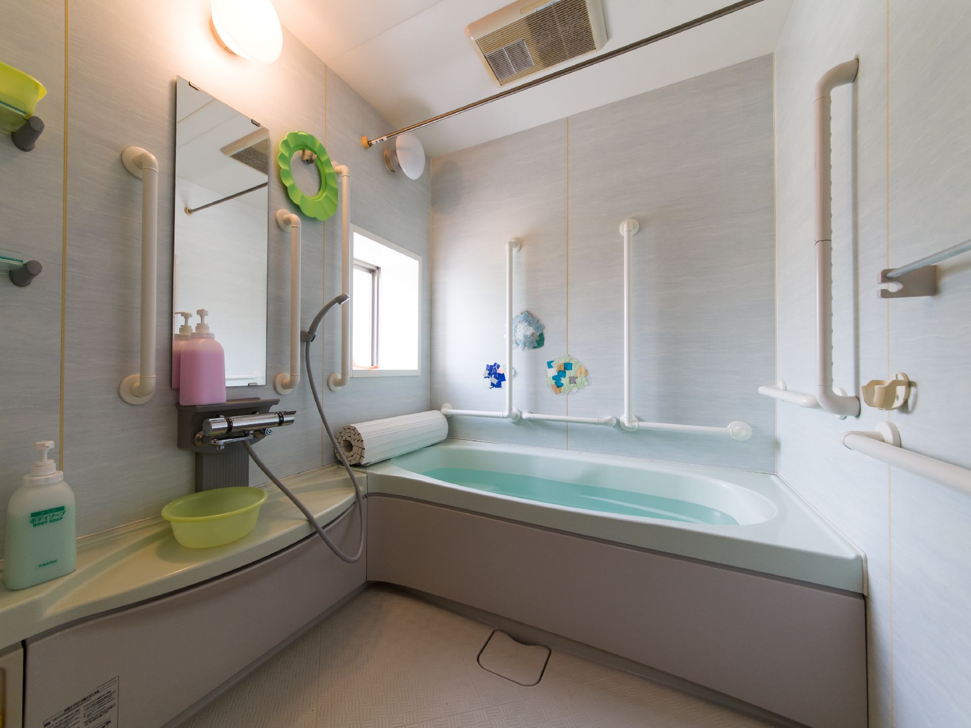 手すりが壁に取り付けてある。鏡の横にシャワーが取り付けてある。お風呂の蓋が浴槽の上に置いてある。ボディソープ、シャンプーなどを置く棚がある。