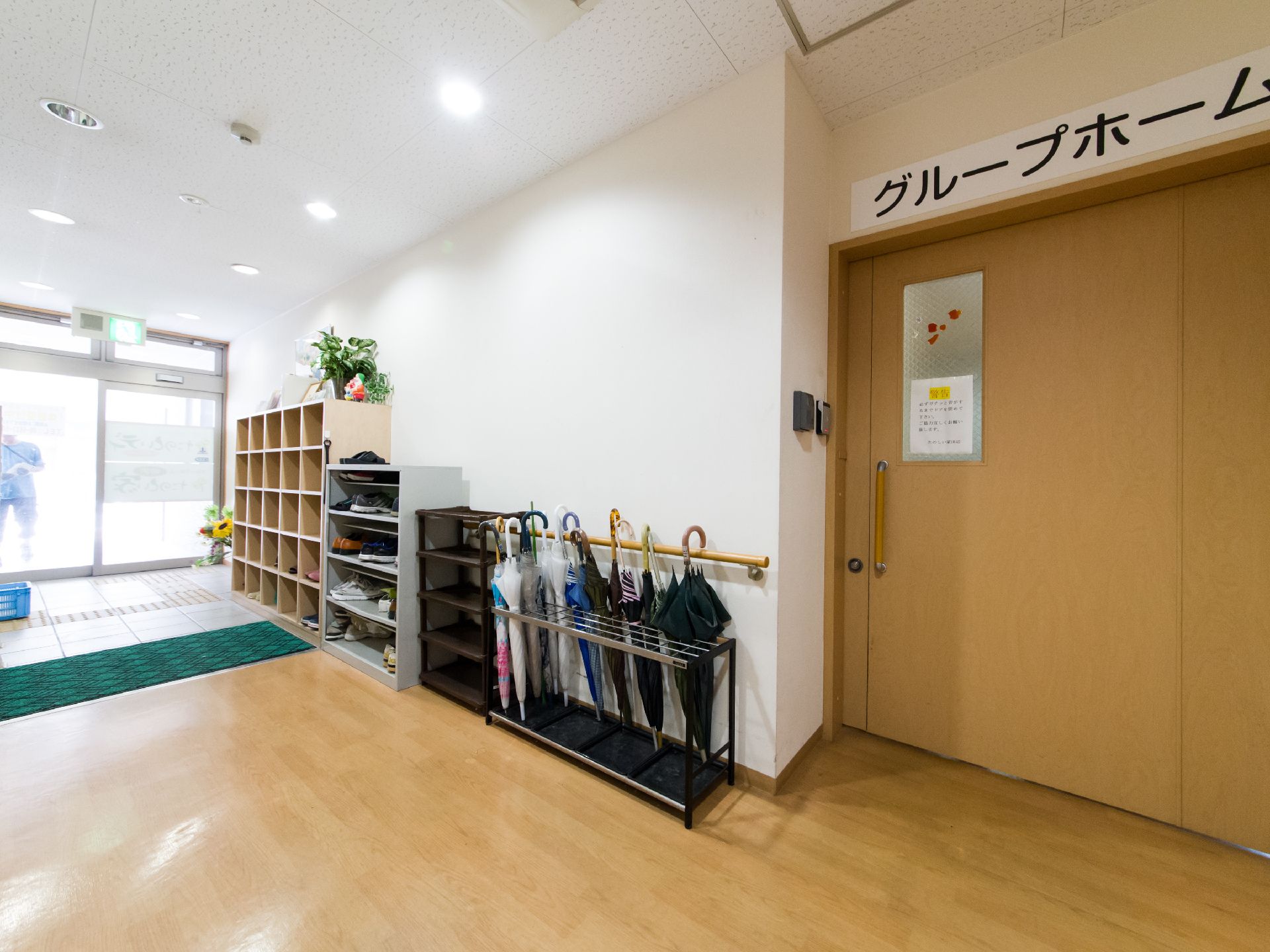 木製とスチール製の靴箱が置いてある。マットが敷いてある。傘立てがあり、傘が数本差してある。靴ベラが靴箱の横のフックにかかっている。