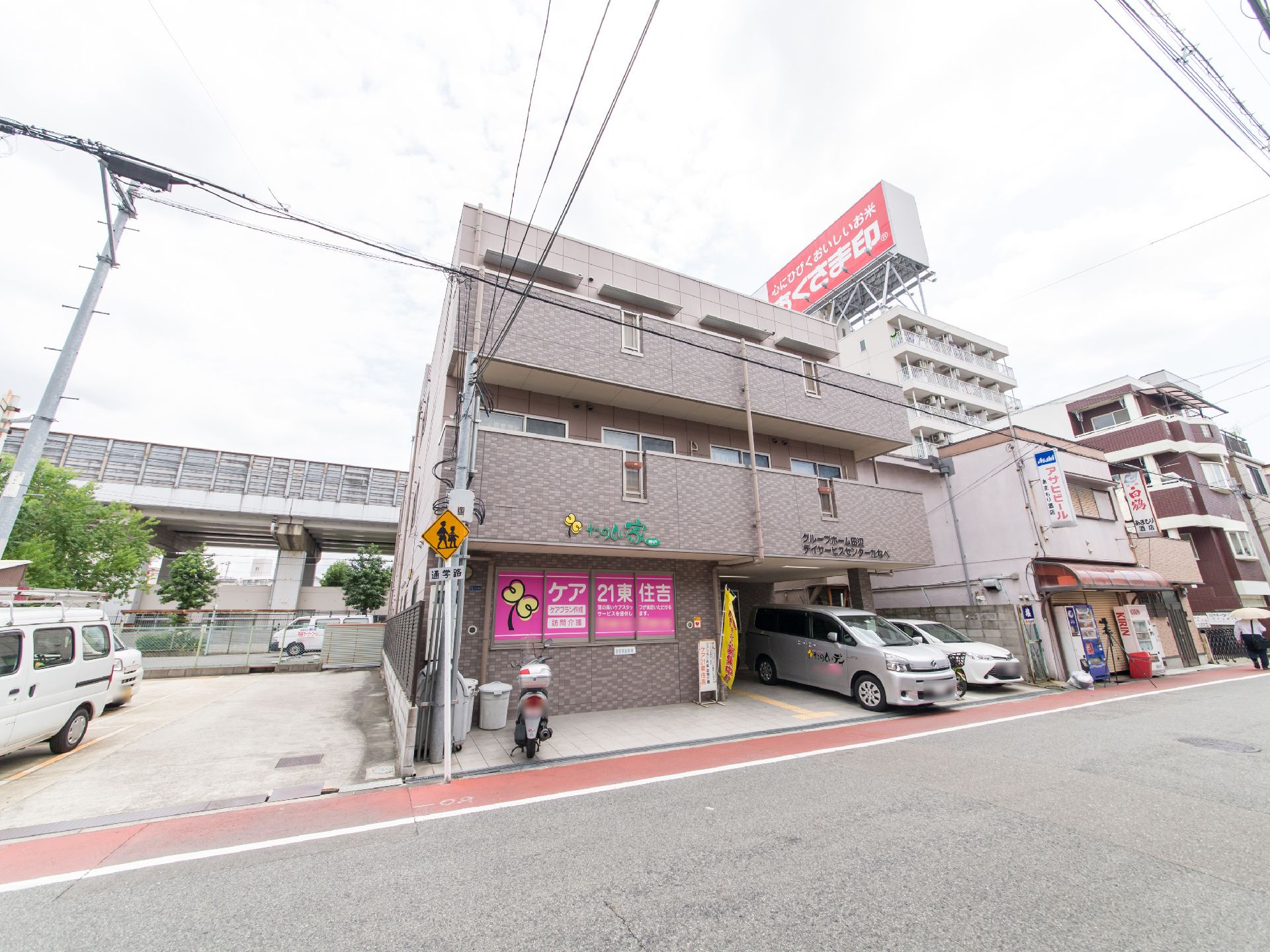 三階建ての施設で、一階部分に駐車場が設けてあり、車が二台停まっている。原付バイクが一台とまっている。黄色の旗が、入り口付近にある。