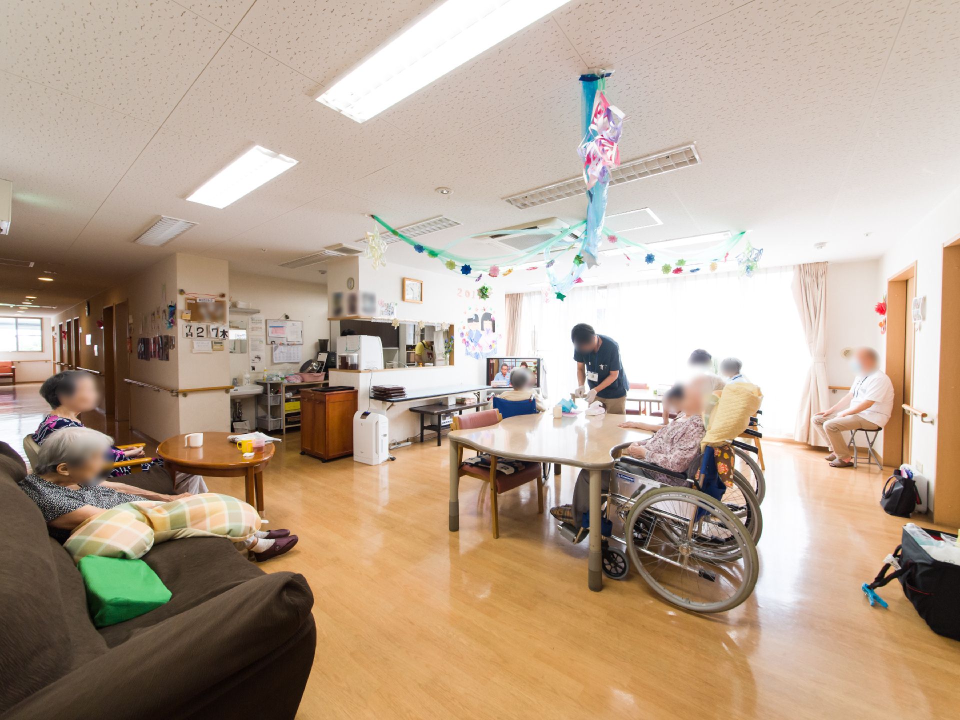 キッチンが見える。テレビがあり、車椅子やソファに腰かけてテレビを見ている入居者がいる。天井から折り紙などで作られた飾りが飾ってある。