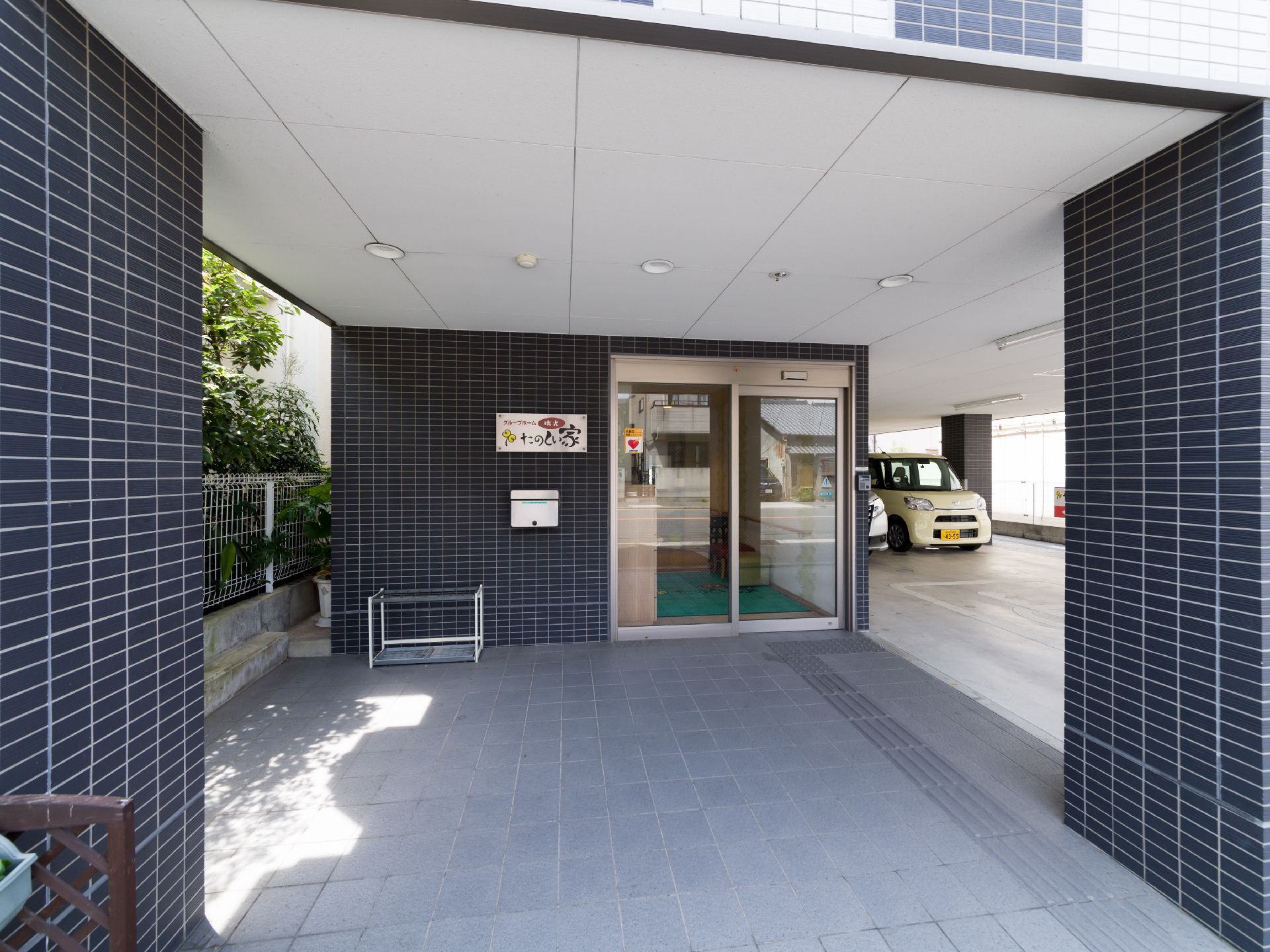 玄関のそばには、傘立てが置いてある。隣の建物との間に白いフェンスが建てられている。玄関は自動ドアである。