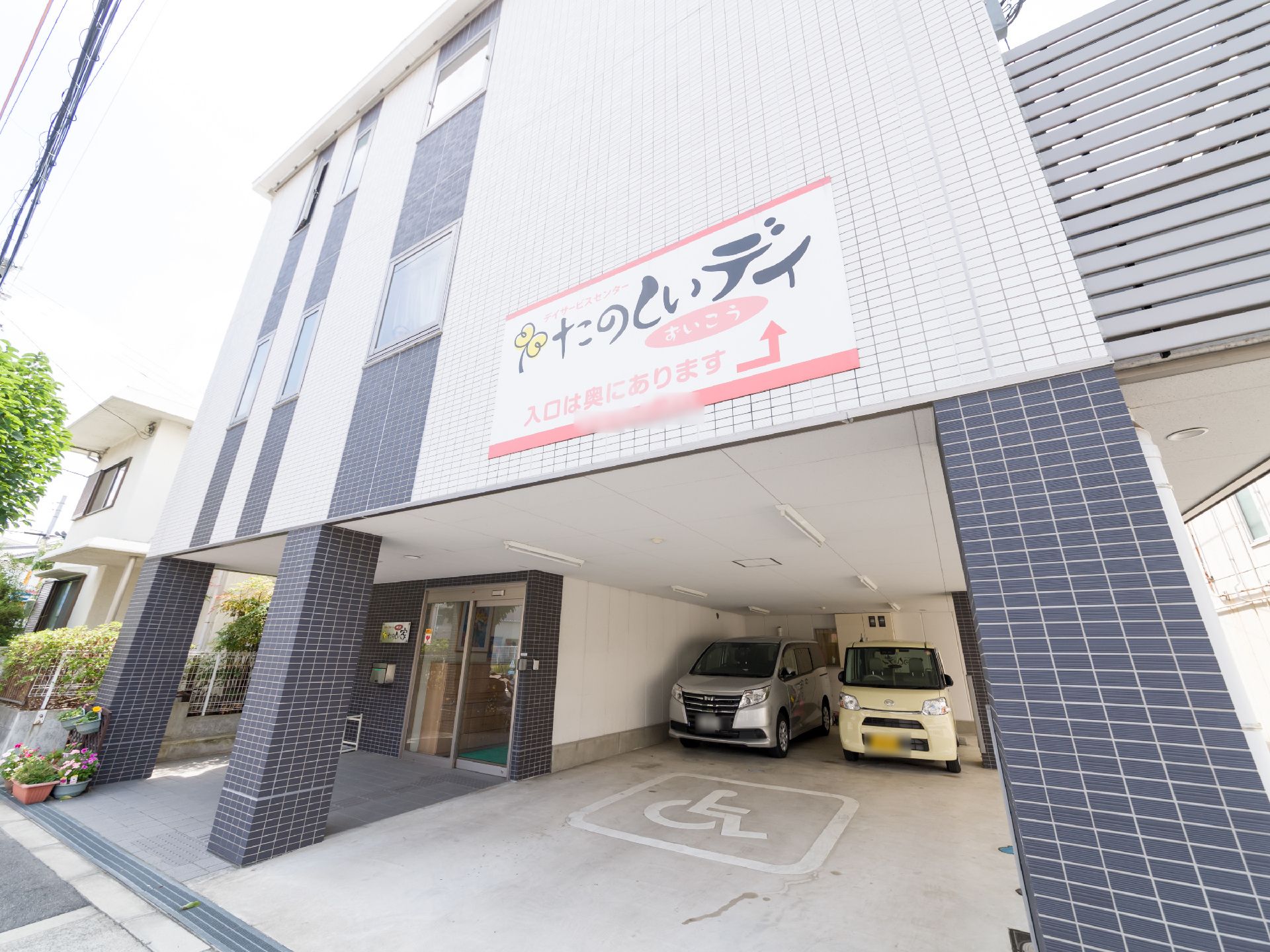 施設の外壁は全体的にタイル調になってる。2階が突き出していて玄関と駐車場の部分が屋根となっている。看板には、デイサービスの入口が表示。