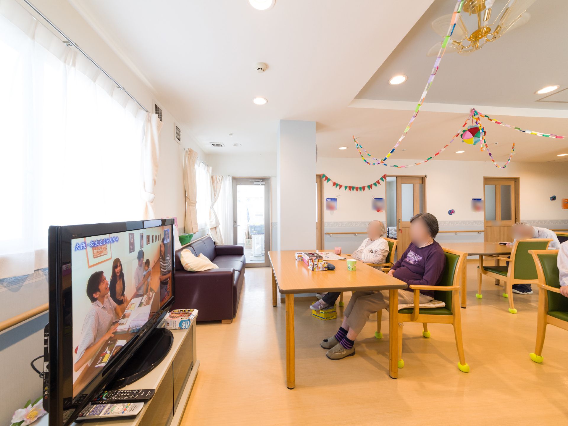 窓のそばにテレビが置いてあり、その横にソファがある。食堂には入居者がいて、椅子に座ってテレビ鑑賞をすることができる。