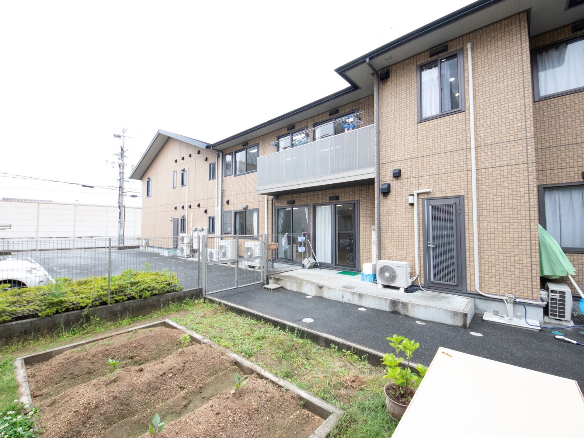 ２階建ての建物で、外壁の色はブラウン。建物前には畑があり、フェンス向こうがわには駐車場が完備されている。