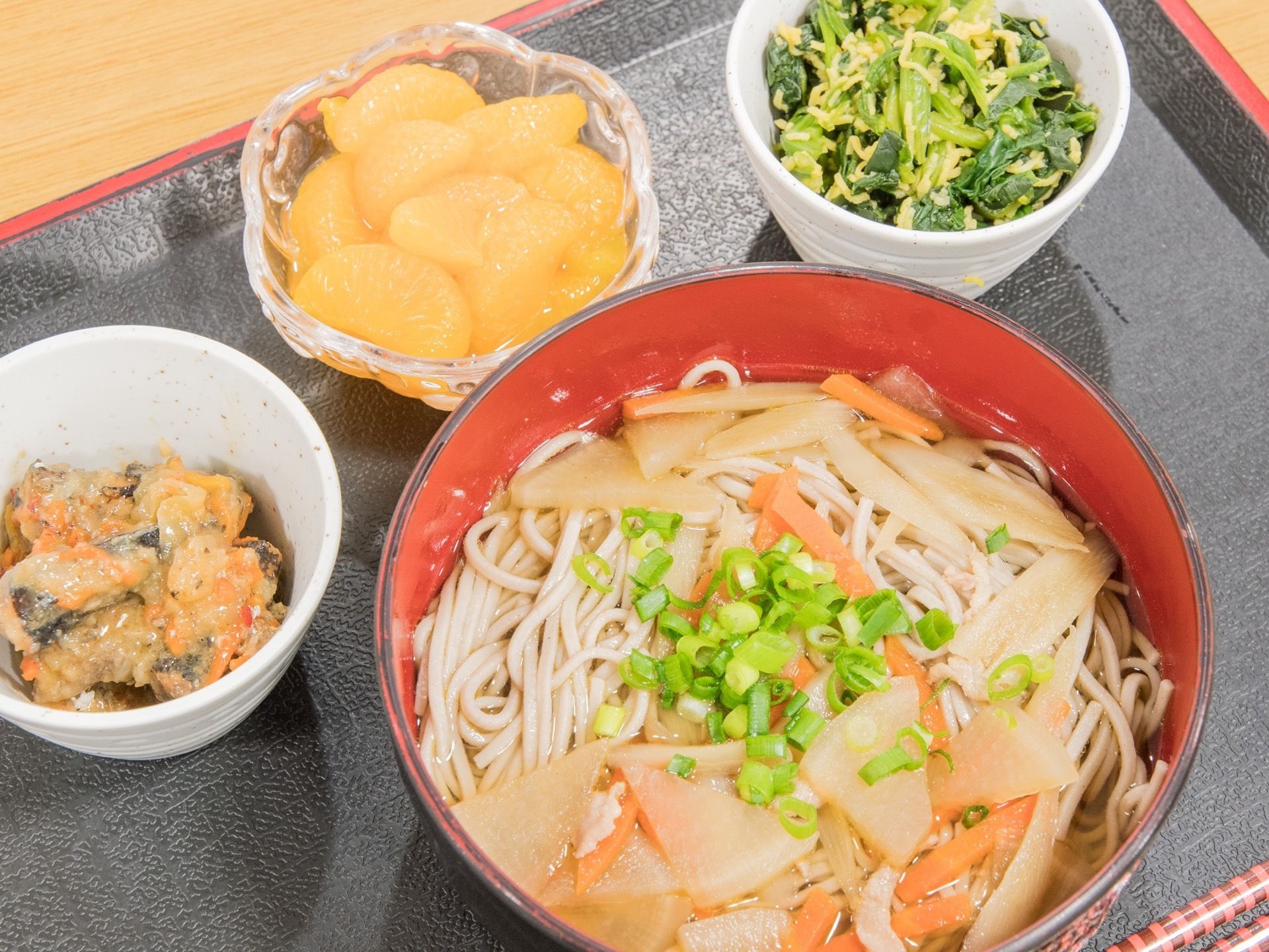 汁物も兼ねたメイン料理の細麺や２品の副菜、デザートの缶詰ミカンが入った小鉢が黒いトレーに並べられている。