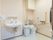 広々としたトイレである。洋式便器の他に洗面台も設置されている。扉付きの収納棚やＬ型手すりが壁に取り付けられている。