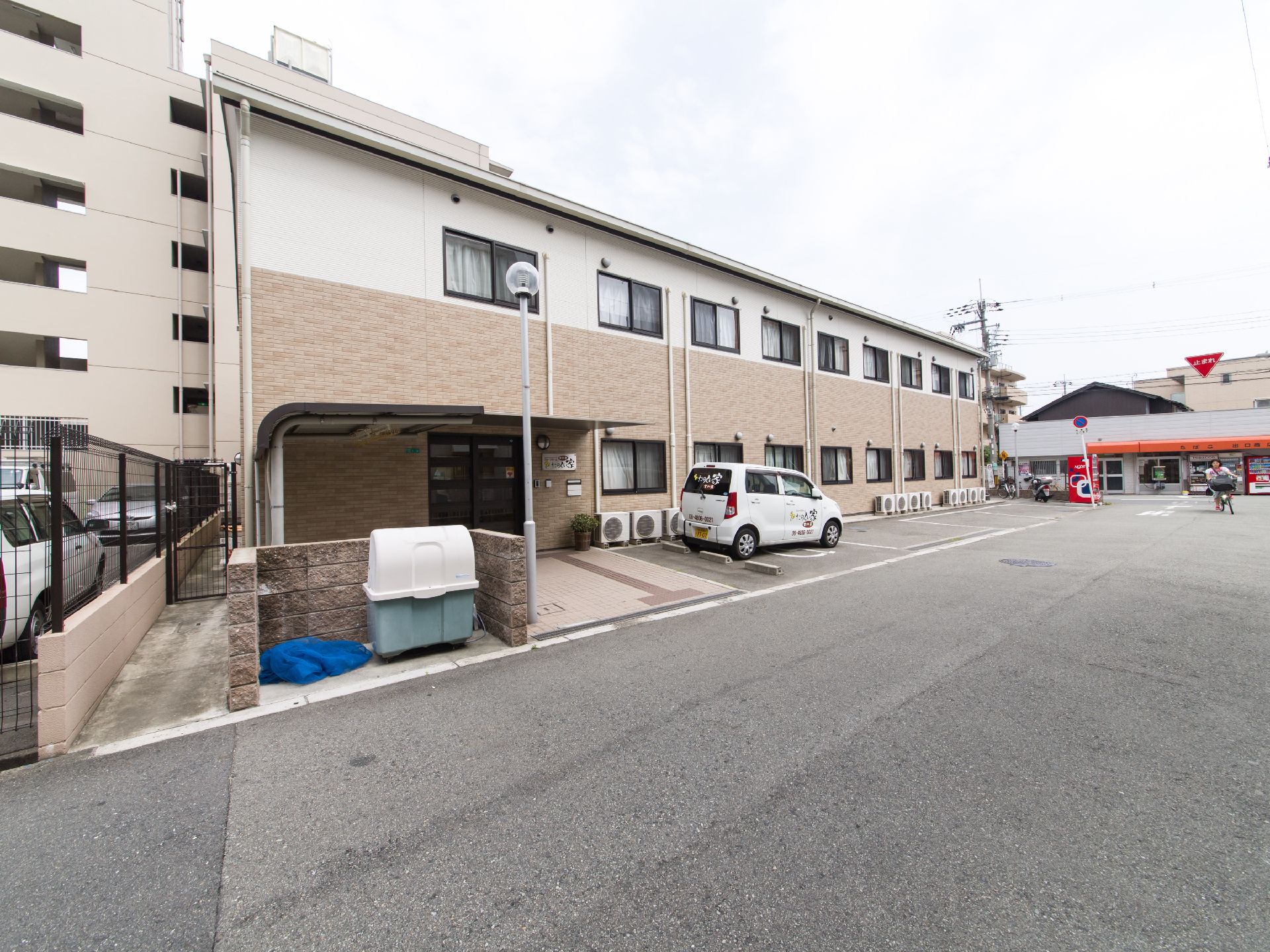 二階建て茶色の建物で、建物の前面に駐車スペースが設けてある。ごみ置き場は塀で囲まれている。玄関そばに観葉植物がある。