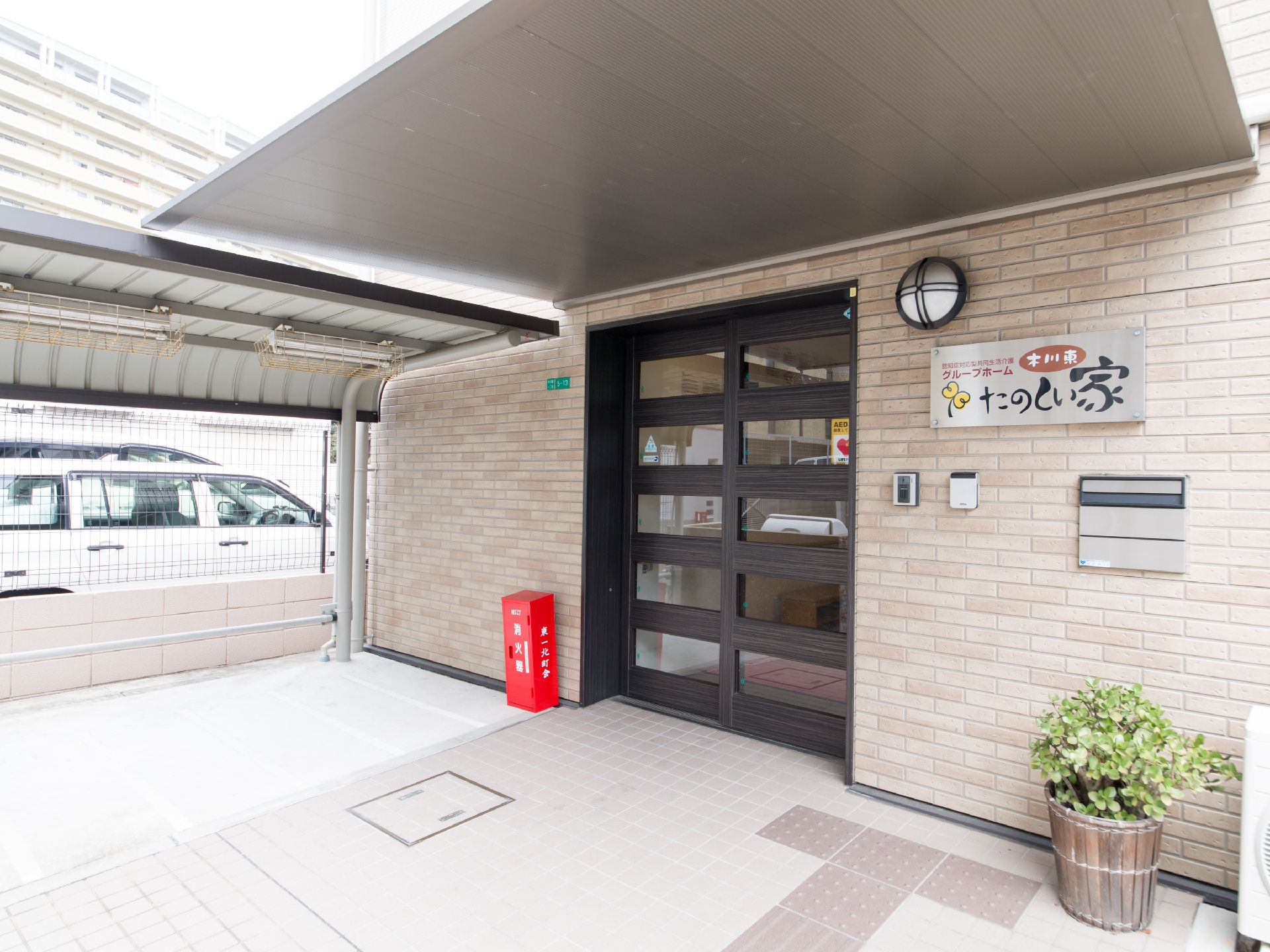 濃い木目調とガラスのツートンになった玄関は落ち着いた印象である。横の外壁はベージュの煉瓦造りになっている。