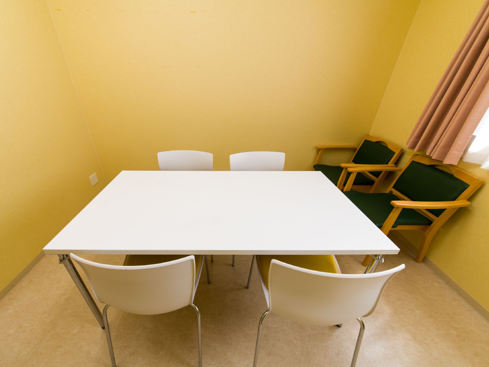 白いテーブルが1つとそれを囲むように四つの白い椅子が並べてある。部屋は、エアコンがとりつけてある。壁に二脚緑色の椅子がある。