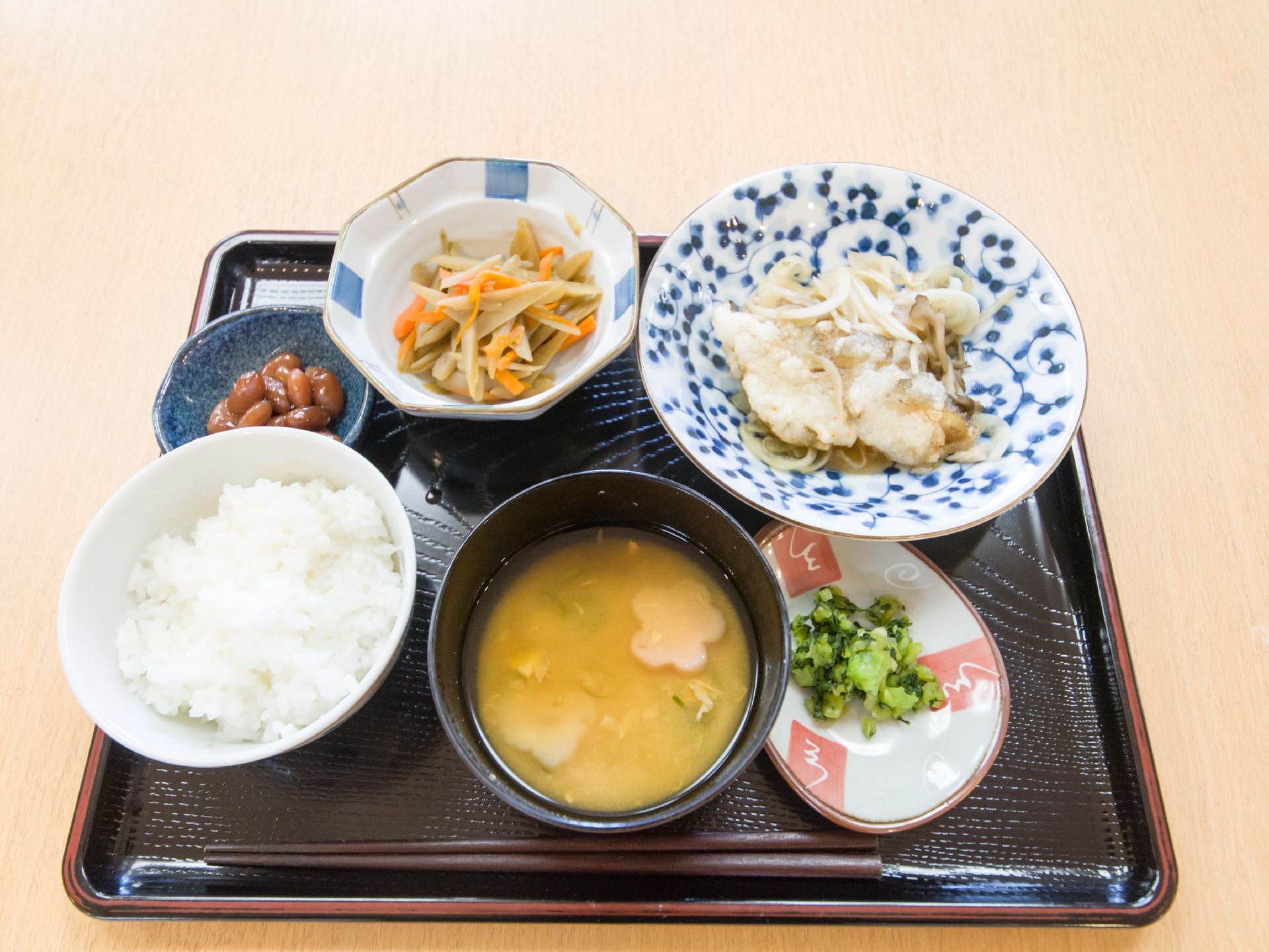黒いお盆の上に食事がおいてある。メイン料理は魚の切り身の揚げ物である。和風の器に盛られて華やか。