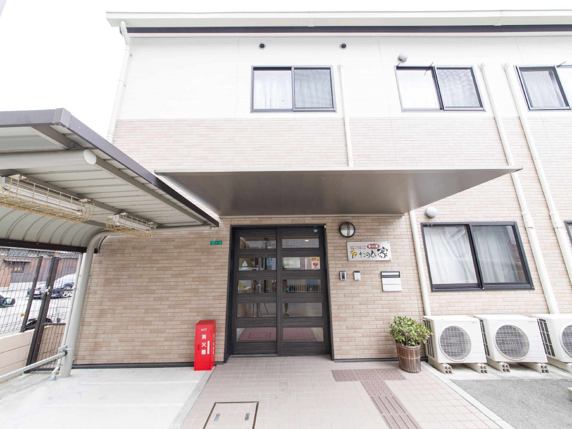 玄関にはかなり大きなひさしが突き出している。横には電気の付いた駐輪場がある。窓の下に室外機が並んでいる。