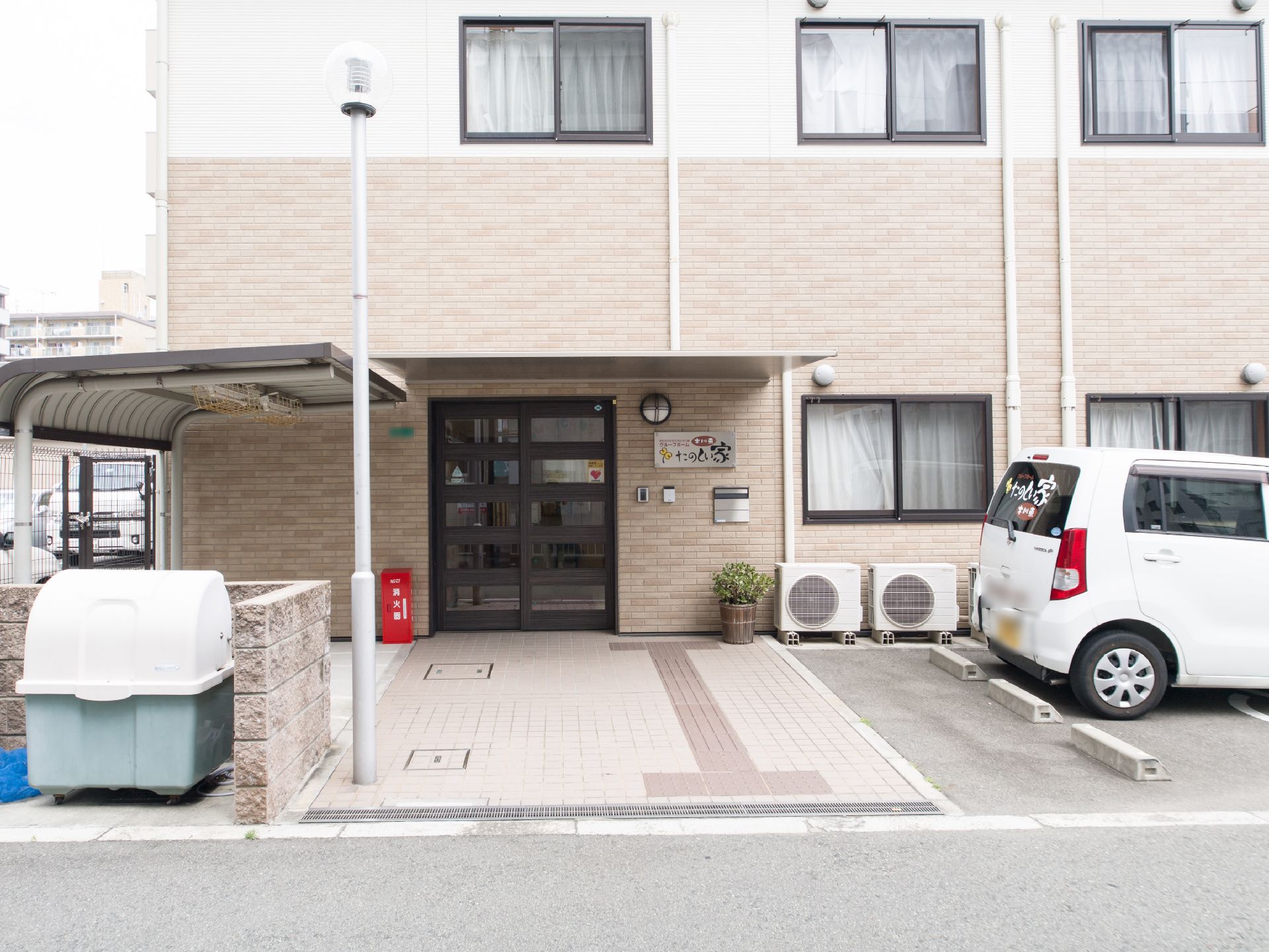 1階2階共にカーテンのかかった窓がある。玄関入口の横には施設名が書かれた看板、インターホン、郵便受け、ポーチライトがある。