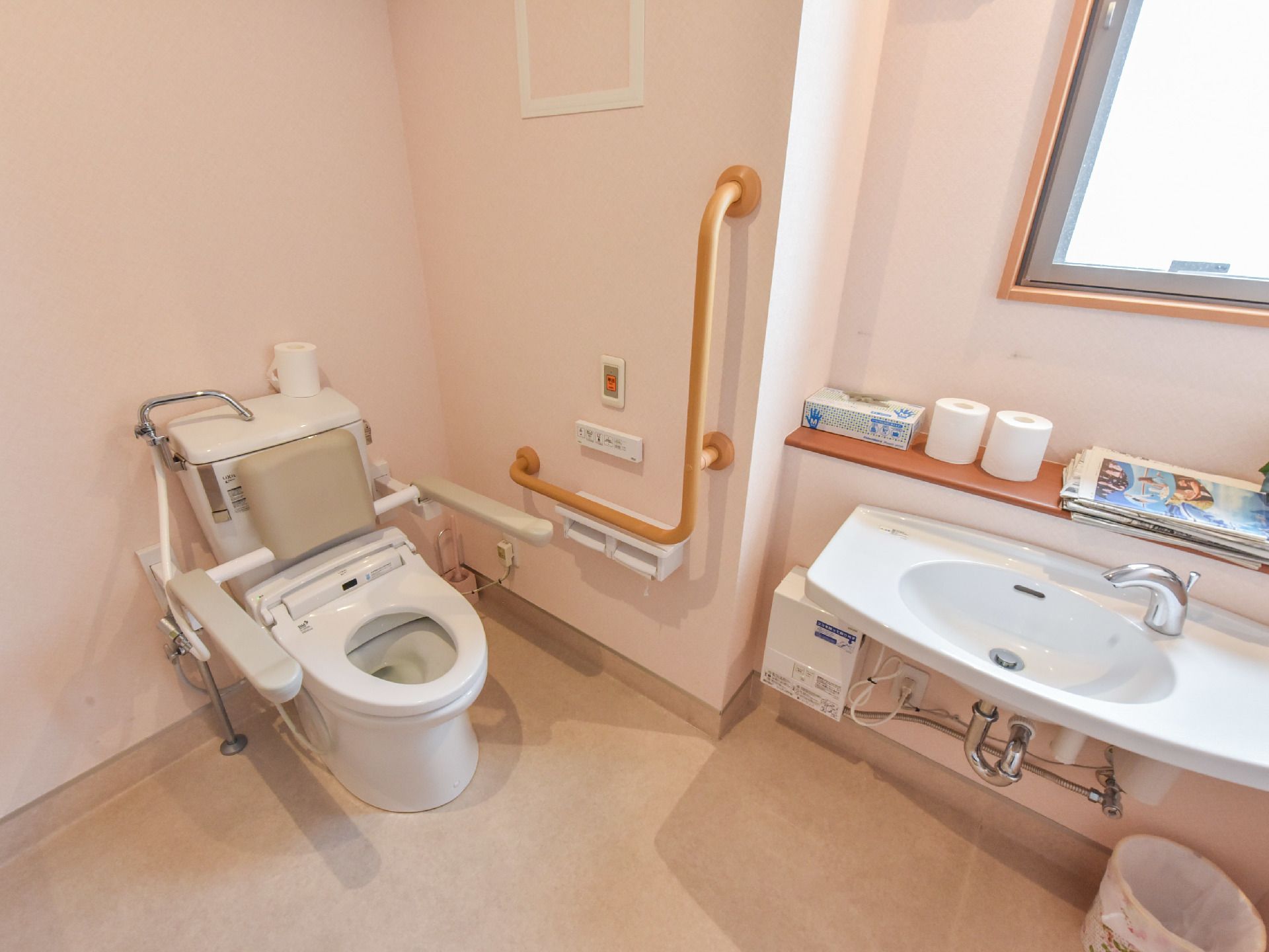 トイレの壁紙は淡いピンク色で床はベージュ色の軟質の素材で踏み心地が良い。幅広の手洗い器で手を洗い易い。