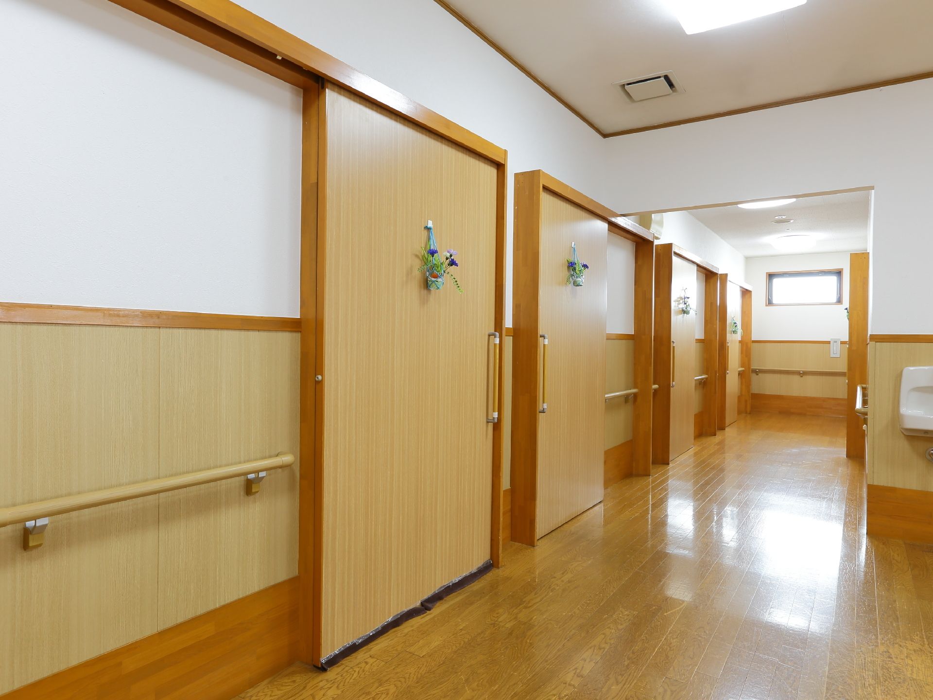 フローリングの廊下に大きな引き戸の扉が並んでいる。扉には小さな花が吊るしてあり優しさを感じる。壁には手すりが付いている。