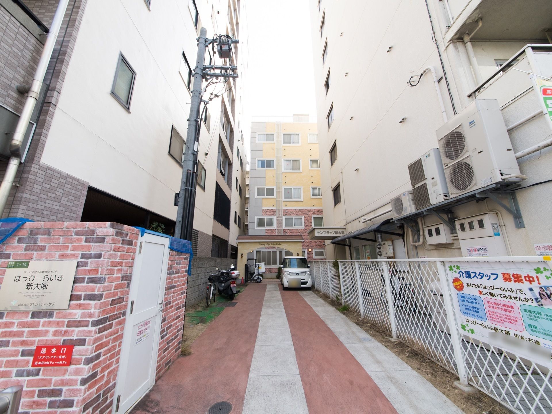 両側に、白色をした大きな建物が建っている。左側にレンガ造りの壁があり、施設名が表示されている。右側にはフェンスがある。