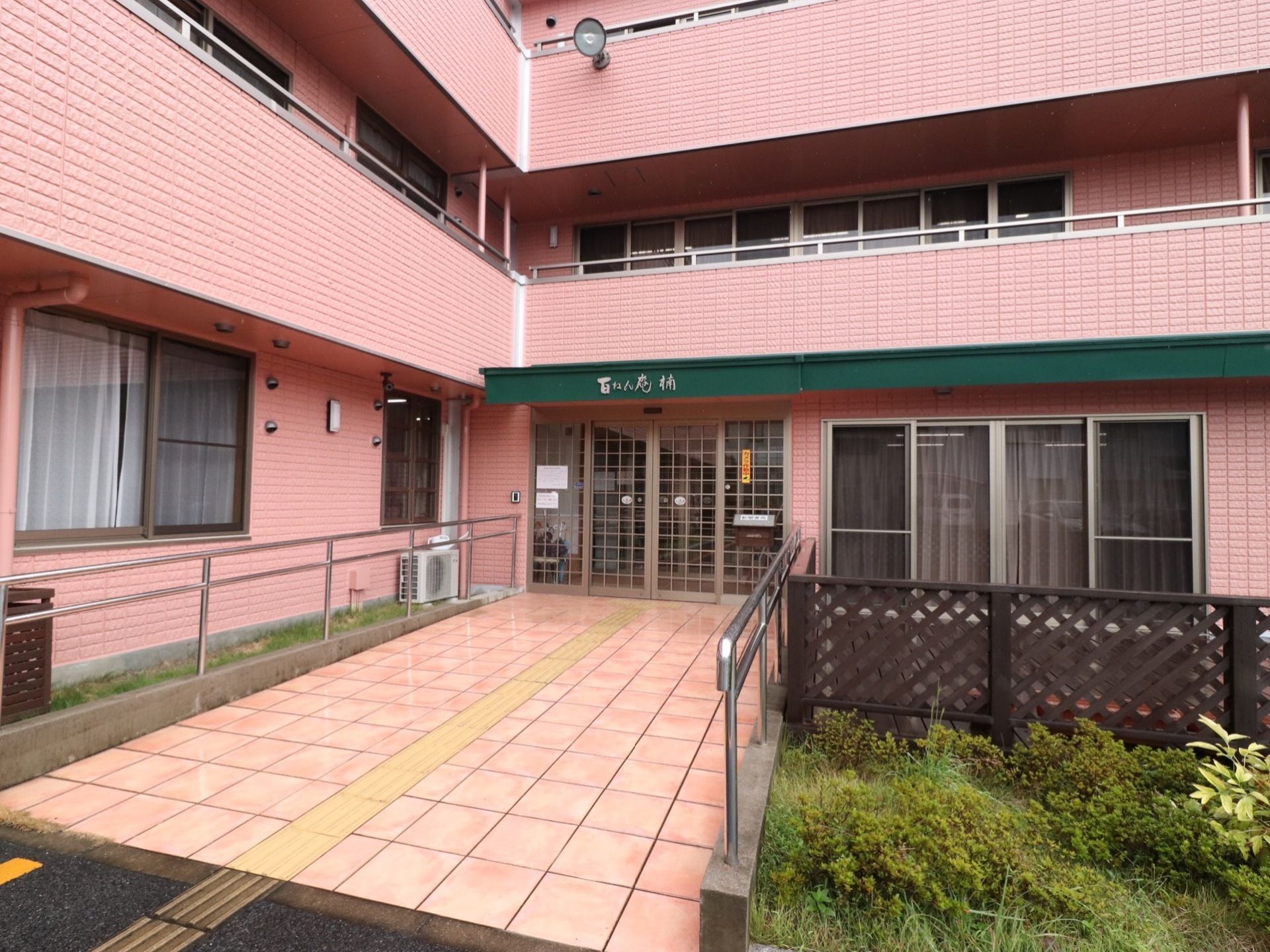 施設の建物の外壁は淡いピンク色で、玄関の上部にはダークグリーンの屋根を設置している。玄関前には植物を植えている。