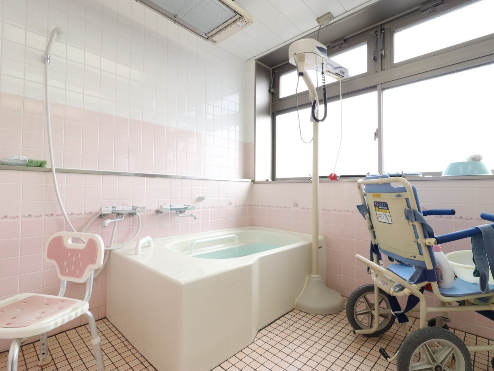 浴室の壁には、白と淡いピンク色のタイルが貼り付けられている。水色のお風呂用車イスの上には、白い洗面器が置いてある。