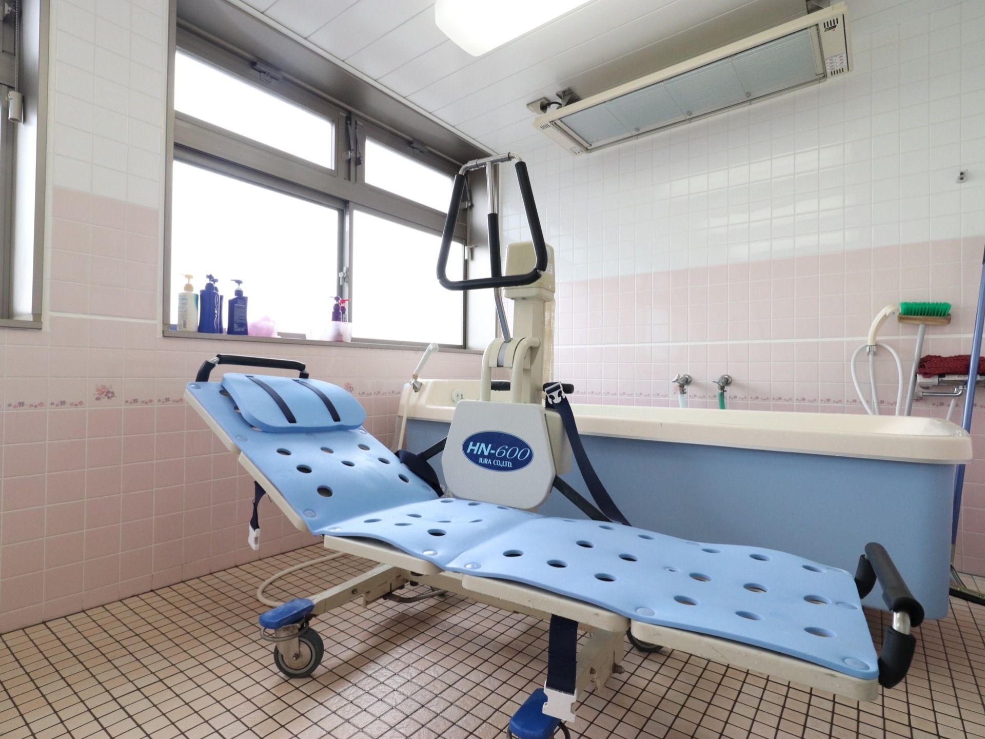大きな介護用の入浴装置は、白とペールブルーが組み合わされた爽やかなデザインである。窓際には、シャンプー等が置いてある。