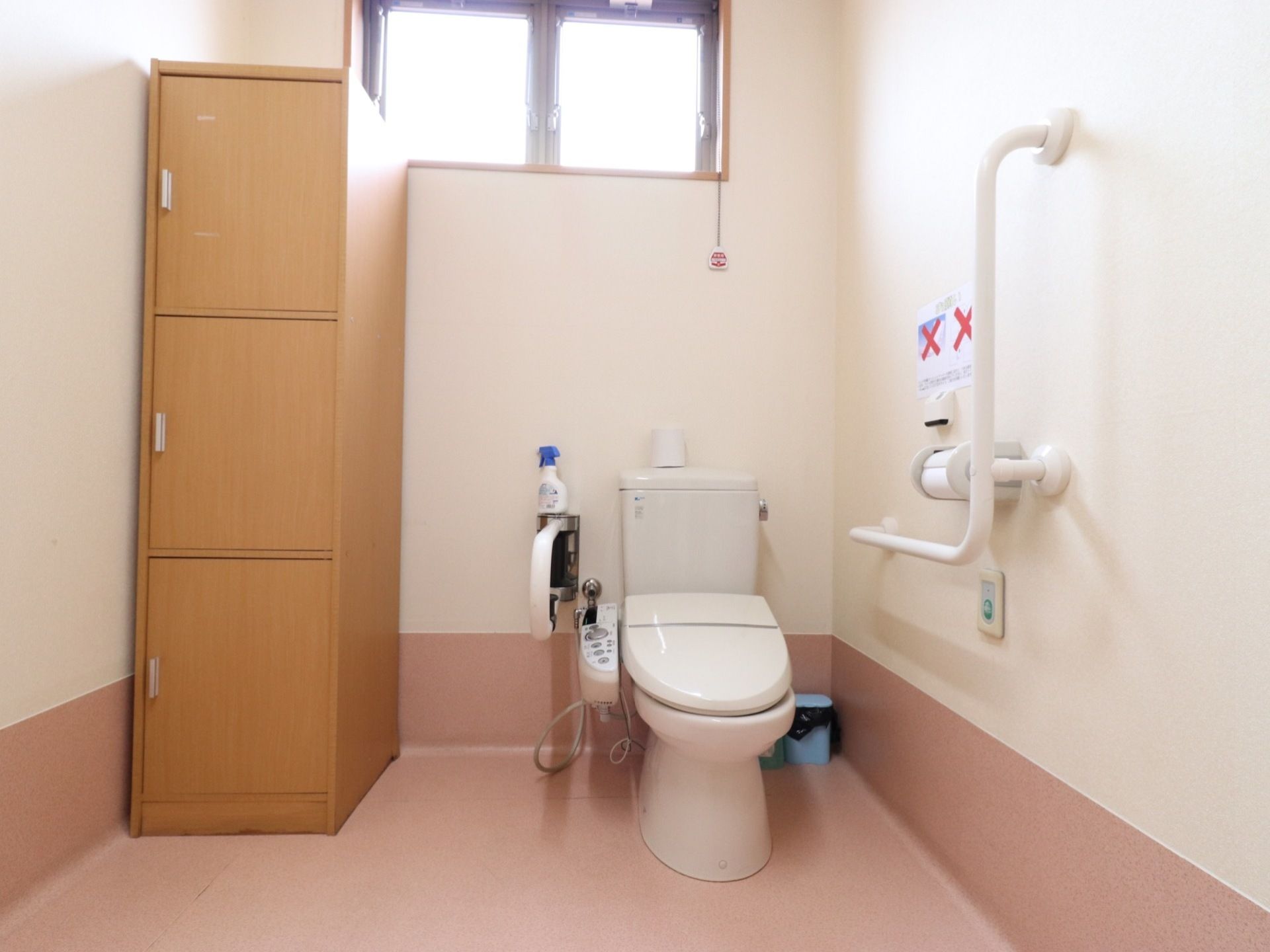 天井近くにある窓から、明るい光が差し込んでいる。トイレ両側の壁には、手すりが設置されている。左端には、収納家具がある。