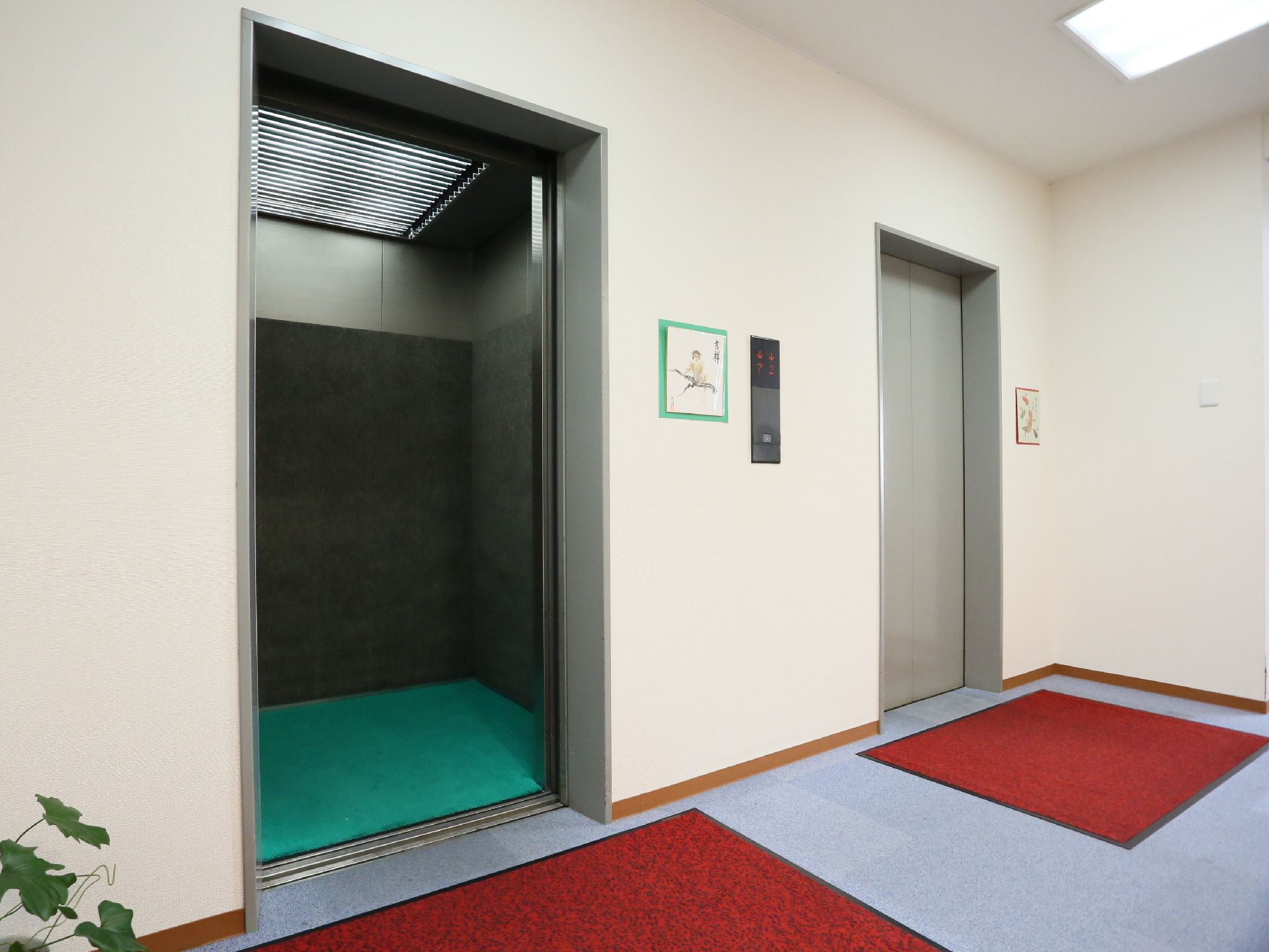 エレベーターが2基並んで設置されており、それぞれのドアの前に赤いマットが敷かれている。壁には操作パネルがついている。