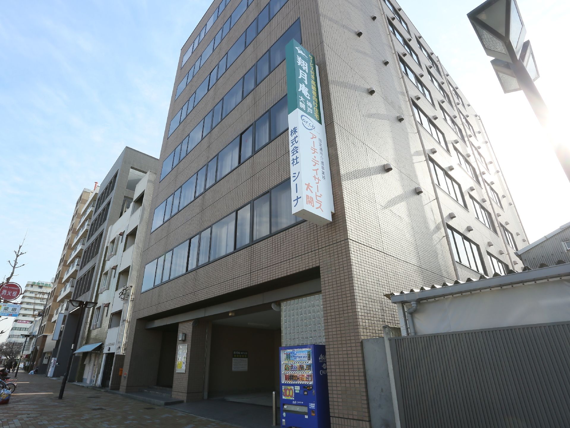 茶色の高い建物になっており、施設名の書かれた看板が取りつけられている。1階の道路に面した場所に自動販売機が設置されている。