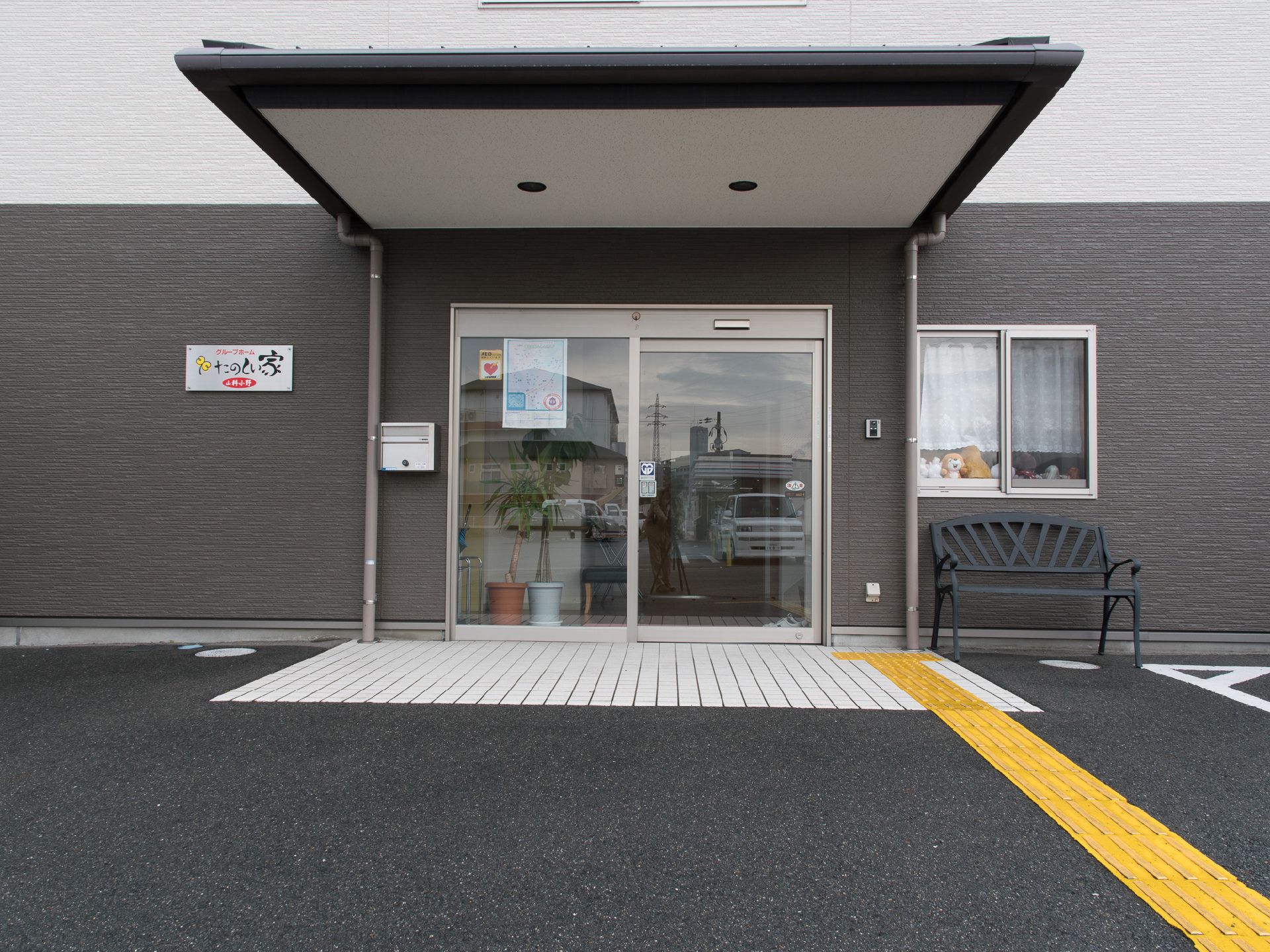 自動ドアの横にポストと施設の表札、インターフォンとベンチが置かれている。玄関前には屋根があり、床には点字ブロックがある。