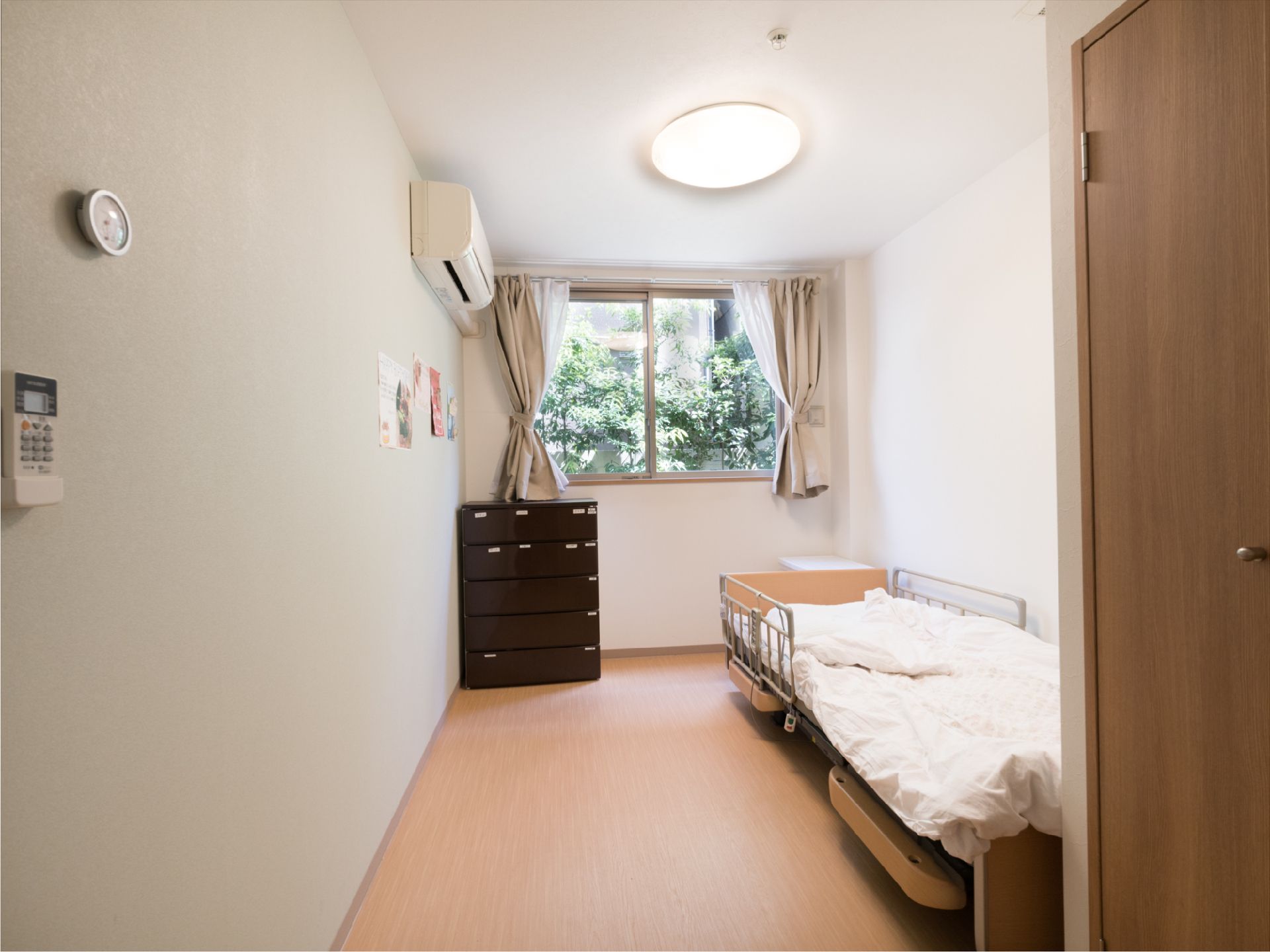 部屋には、電動ベッドが一台置いてある。壁際にチェストがある。腰高窓にはカーテンがあり、庭の木々が見える。
