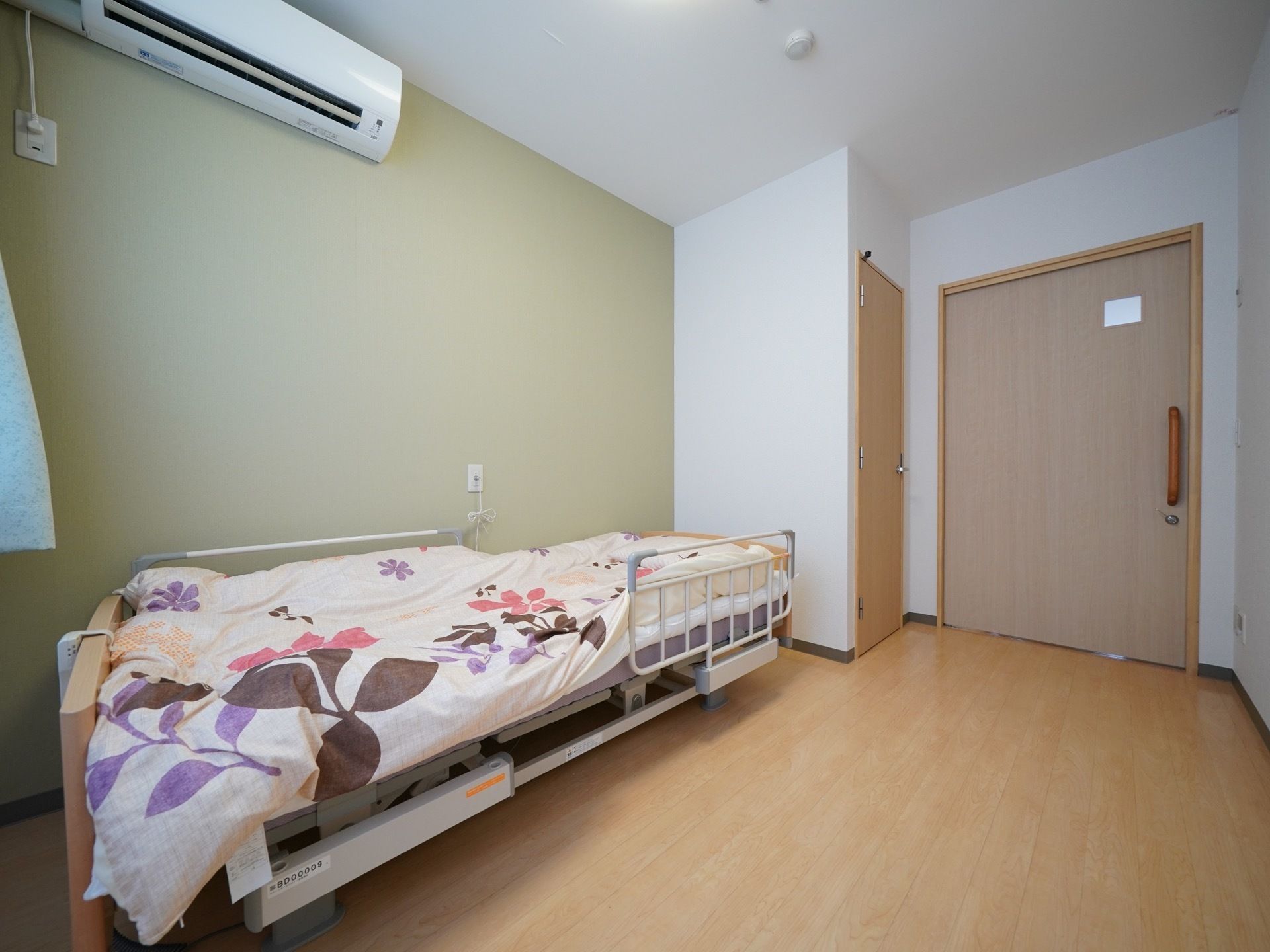 明るい木目調のフローリング部屋である。壁片面が淡いグリーンである。エアコン、介護専用ベッドが完備されている。
