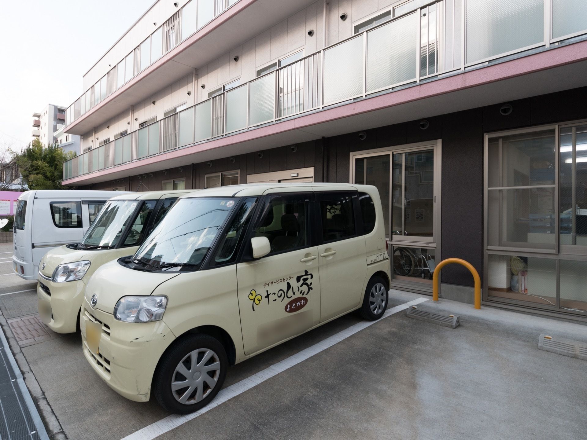 施設は３階建てで横に広い作りである。建物のすぐ側は車の駐車スペースになっている。数台の車が停められている。