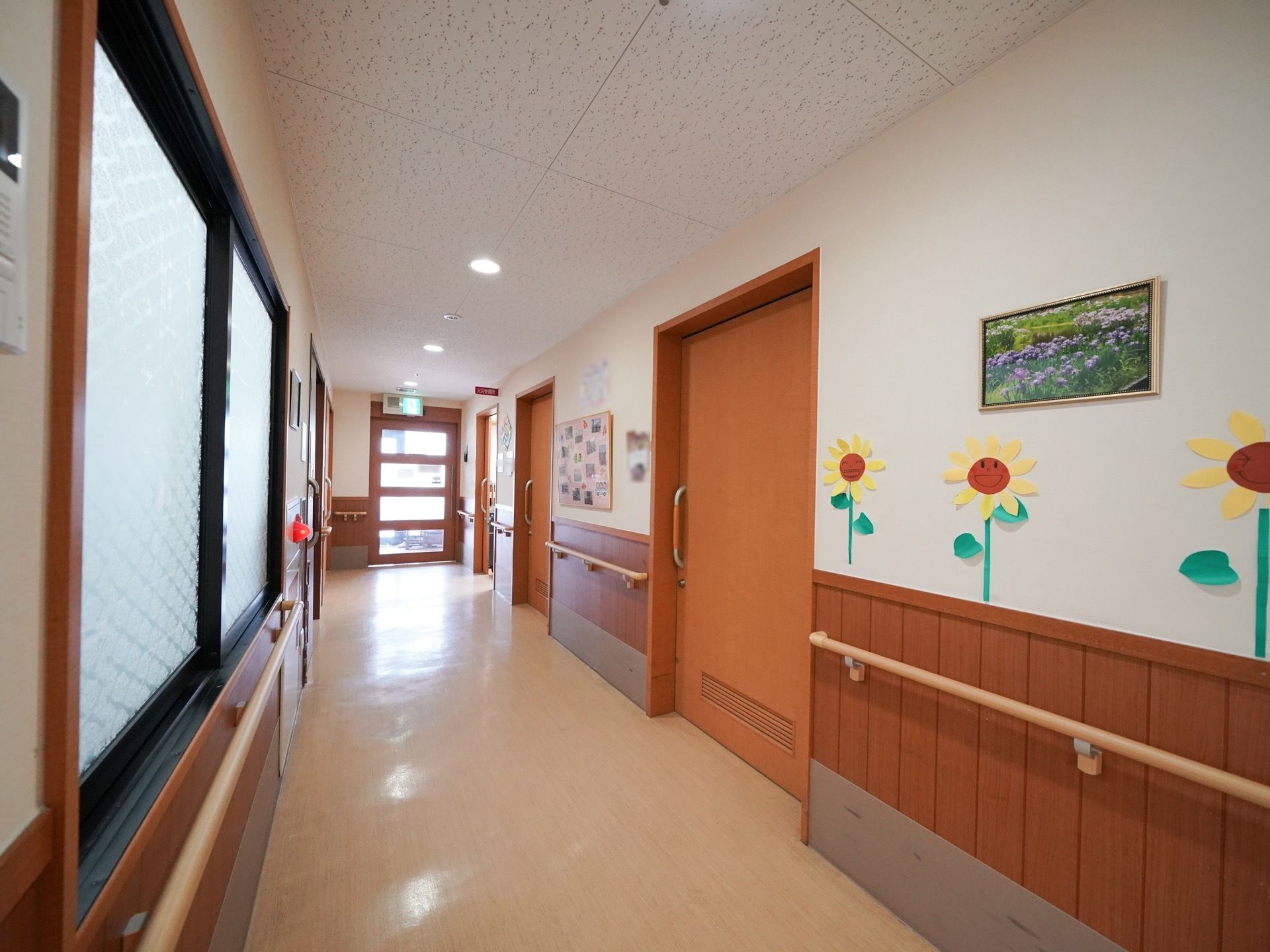 暖色系の床や壁に囲まれた通路。壁には手すりが設置されている。ドアや窓が多数あり、日差しが差し込んでいる。