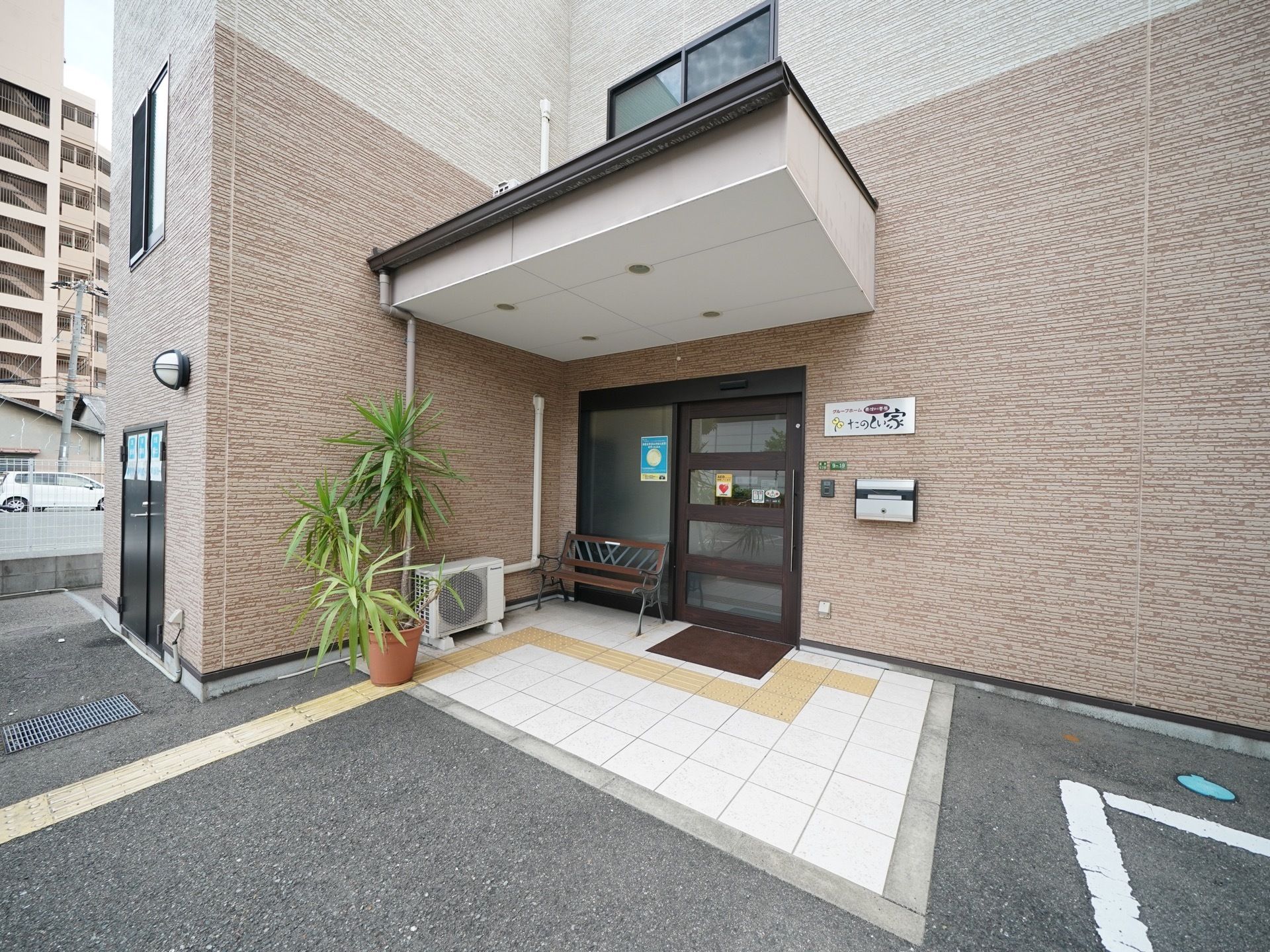 クリーム色と茶色の壁の建物に、濃い茶色の玄関のドアがあり、玄関の横には同じく濃い茶色のベンチがある。建物の前には駐車場がある。