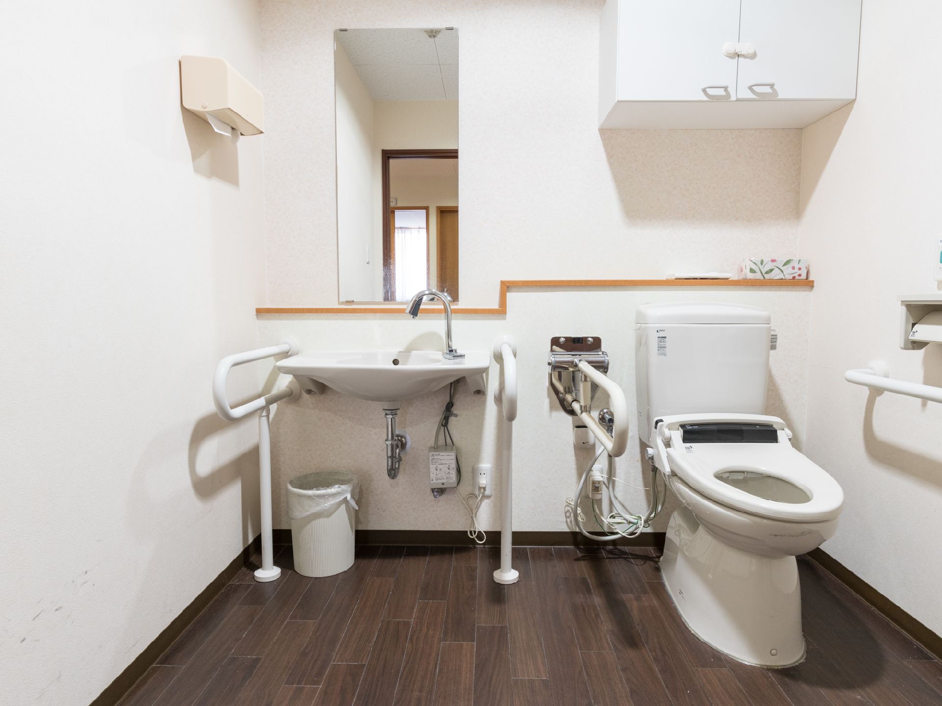 白い壁で囲まれている。画像左側には洗面台が設置され、右側にはトイレがある。トイレの上にはキャビネットが取り付けられている。