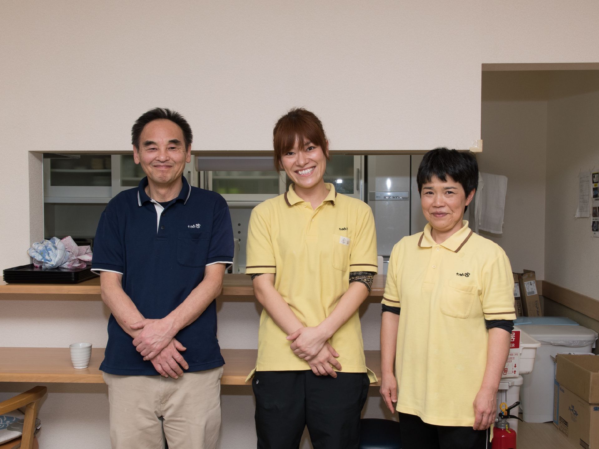 3人のスタッフがにこやかに写っている写真。左端は男性で真ん中と右端は女性スタッフ。背後には食事を提供する調理場が見える。