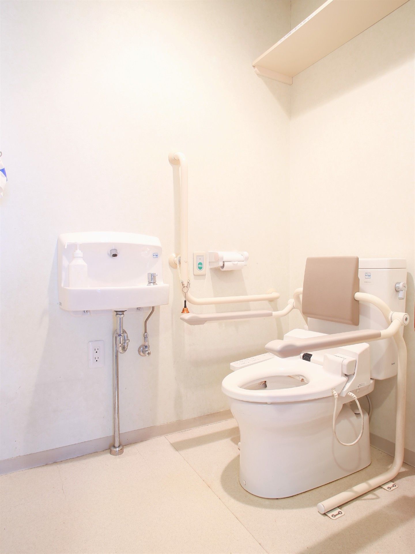 トイレの両側に手すりと背もたれがついている。壁にはL字の手すりと、ミニ洗面台が取り付けられている。呼び出しボタンが設置されている。