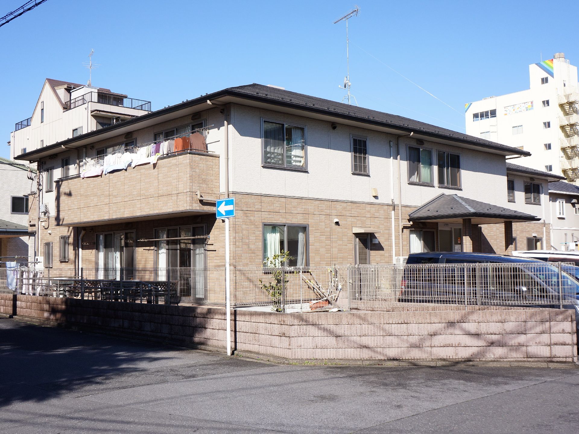 1階の庭と2階ベランダに洗濯物が干されている。2階では布団も干されている。玄関前の駐車場には車が3台ならんでいる。