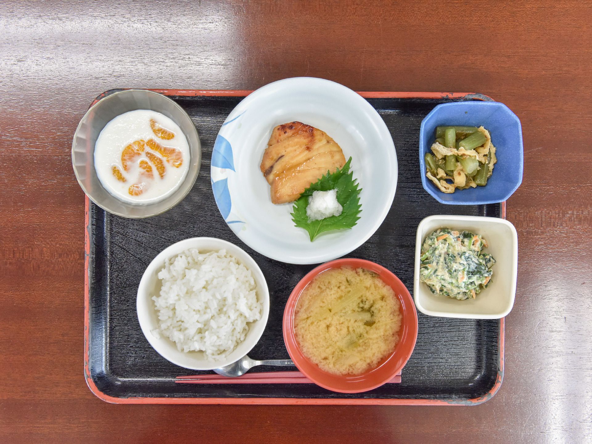 お盆の上にご飯、みそ汁、メイン料理の魚の塩焼き、小鉢2個、デザートが乗っている。お箸とスプーンがセットしてある。